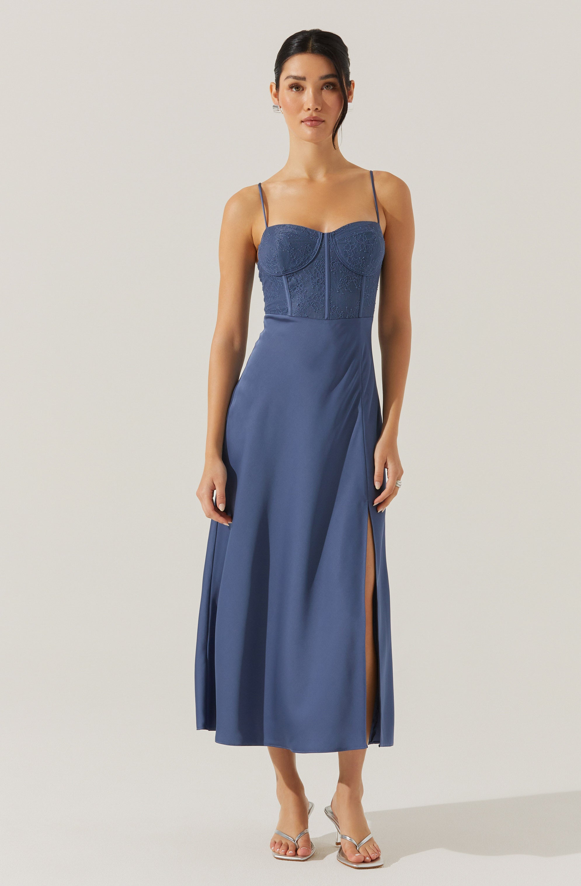 A woman is wearing a sleeveless, fitted blue dress with thin spaghetti straps. The top portion features a lace or embroidered pattern, while the skirt is made of smooth, flowing fabric with a high slit on one side that reveals her leg. She is wearing open-toe, strappy heels.