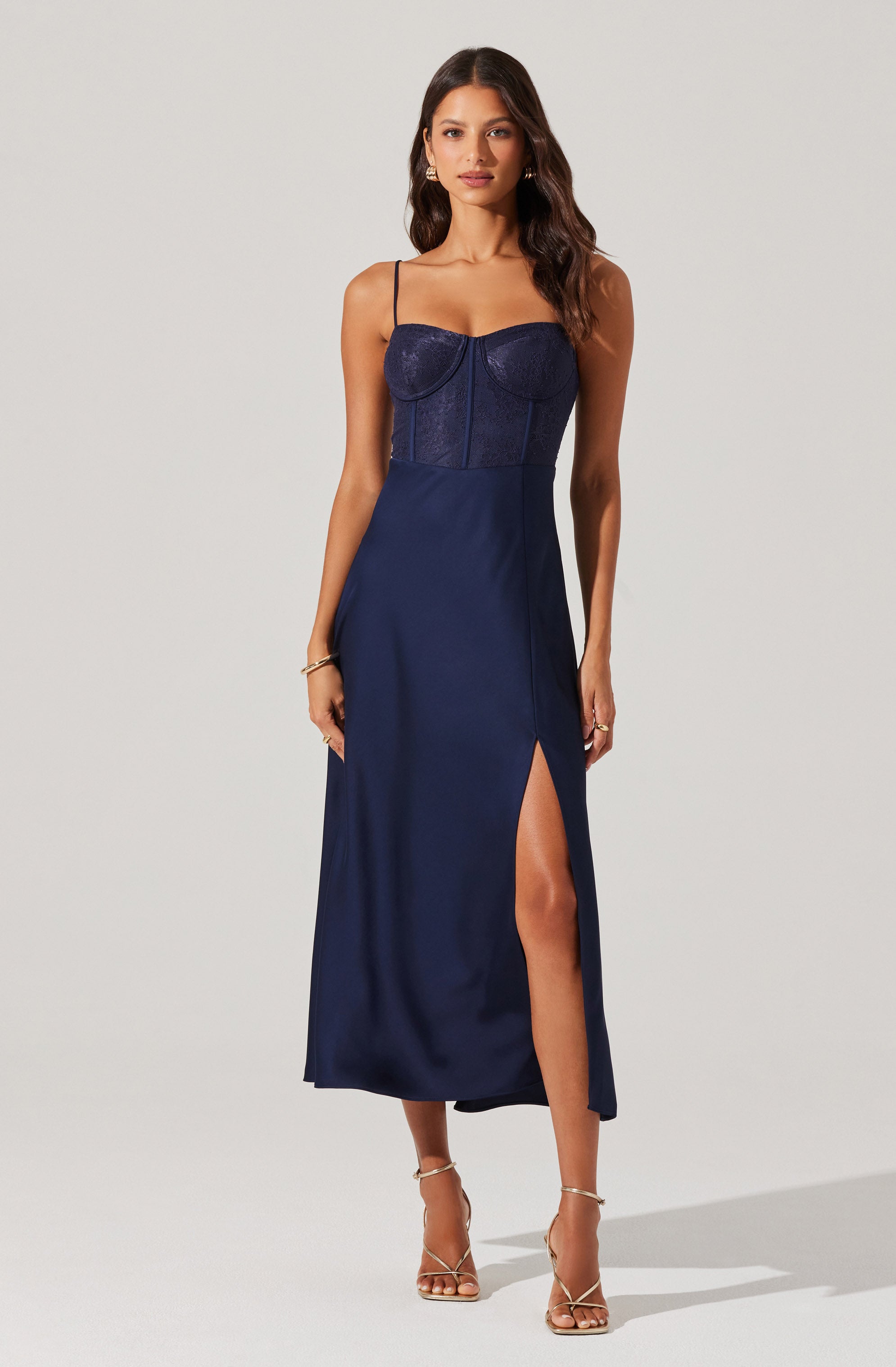 A woman is wearing a navy blue dress with a fitted, strapless bodice that has a sweetheart neckline and thin spaghetti straps. The bodice appears to be made of lace or a textured fabric, and the skirt is flowing with a high slit on one side. The woman is also wearing gold strappy high-heeled sandals.