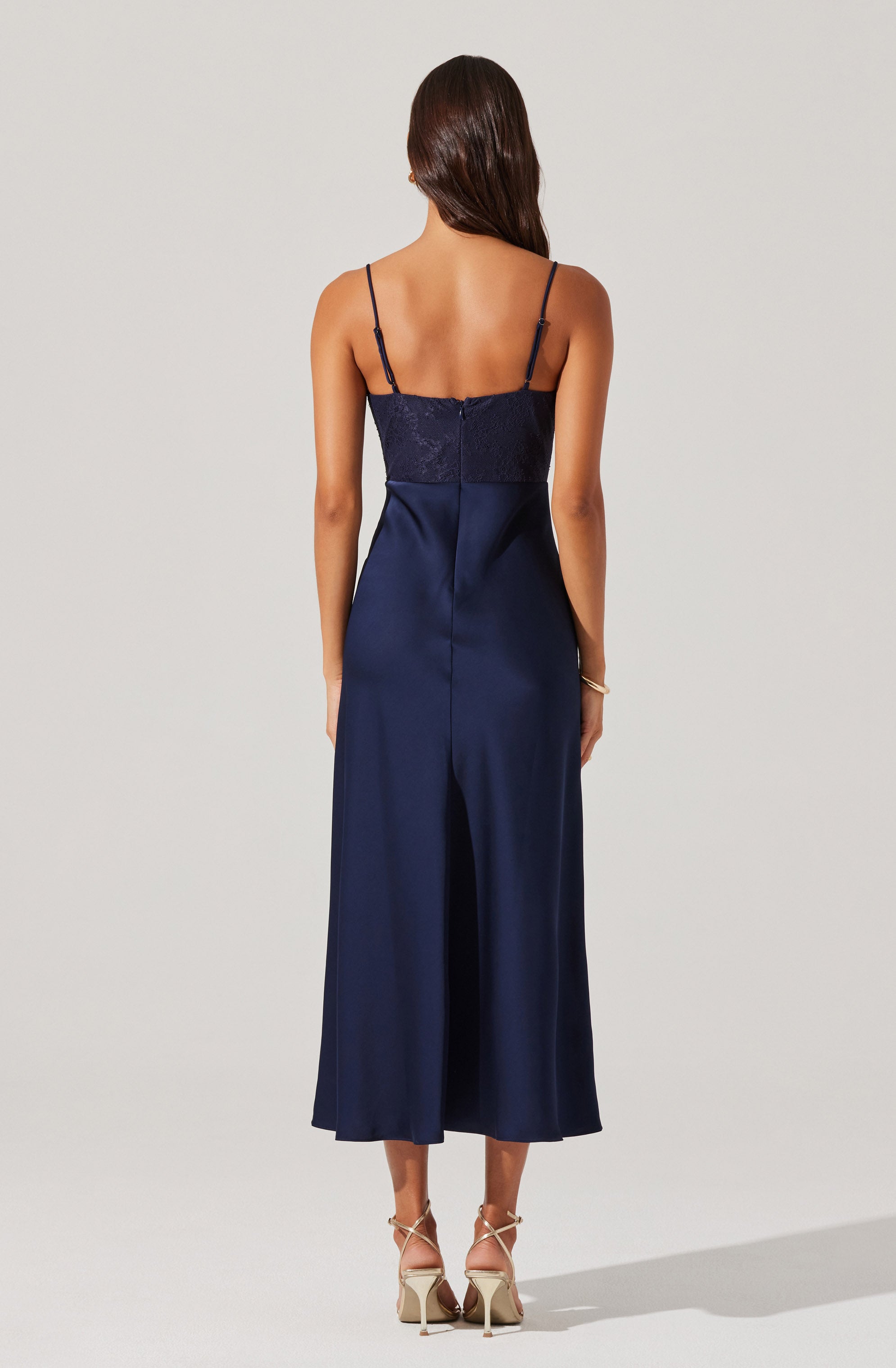 A woman is wearing a navy blue satin dress with a fitted bodice featuring a delicate lace overlay and thin spaghetti straps. The dress has a straight neckline at the front and a long, flowing skirt that reaches her ankles.
