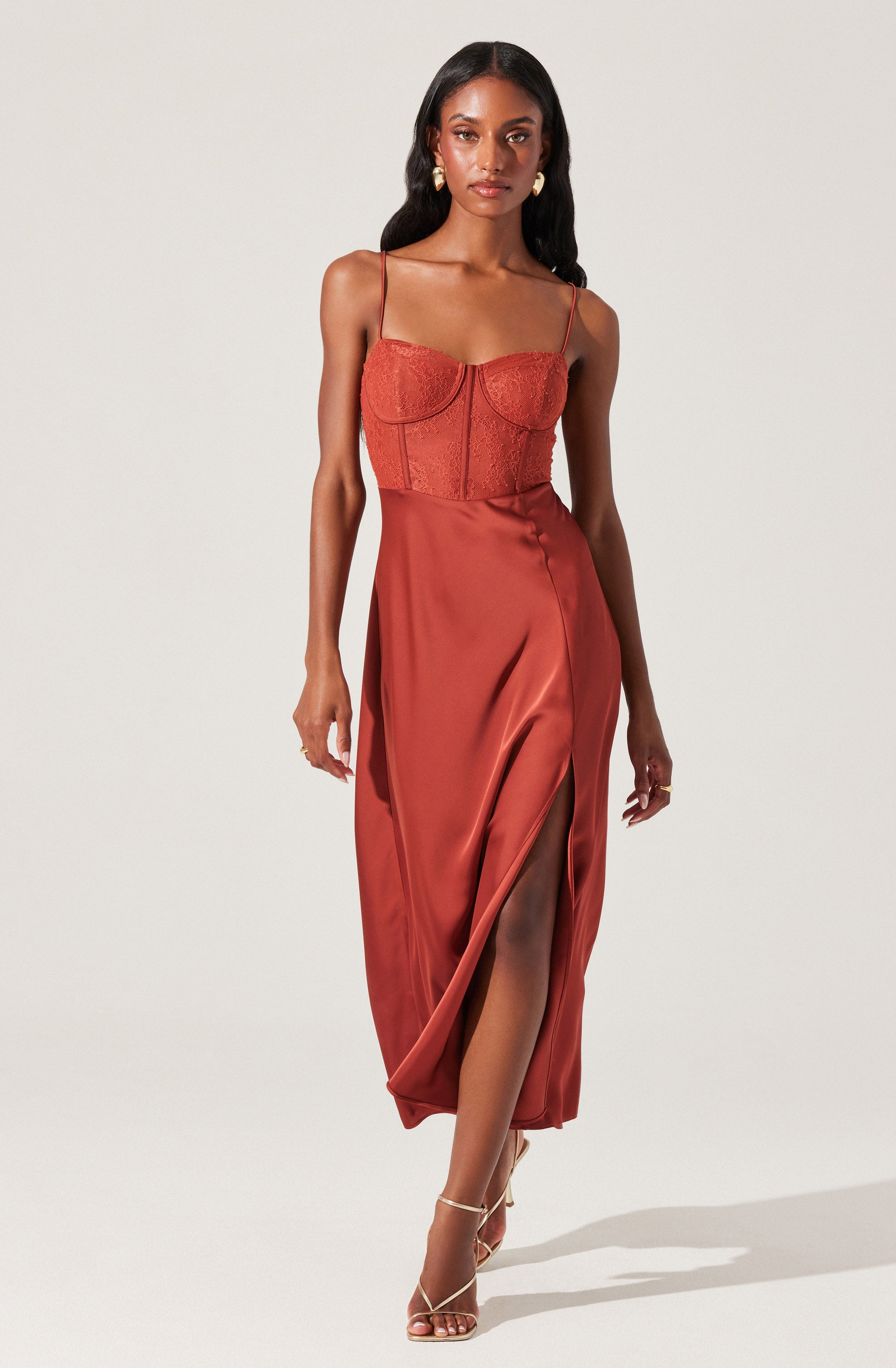 A woman wearing a rust-colored, sleeveless dress with thin spaghetti straps. The dress features a fitted, textured bodice and a flowing satin skirt with a high slit revealing her leg. She is also wearing strappy high-heeled sandals.