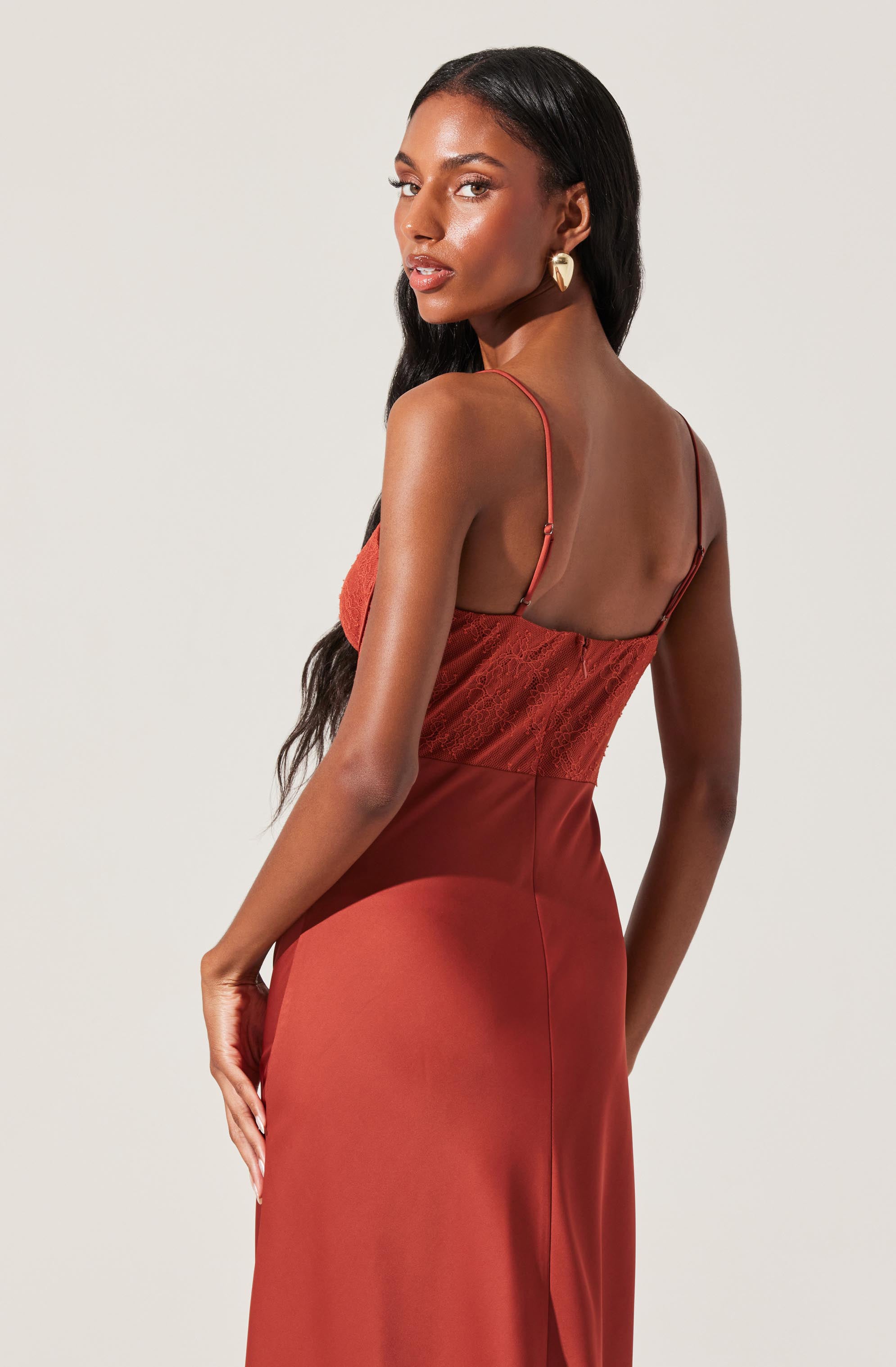 A woman is wearing a sleeveless, rust-colored satin dress with spaghetti straps. The top part of the dress features a lace or textured fabric, while the skirt is smooth and shiny. She accessorizes with large, heart-shaped earrings.