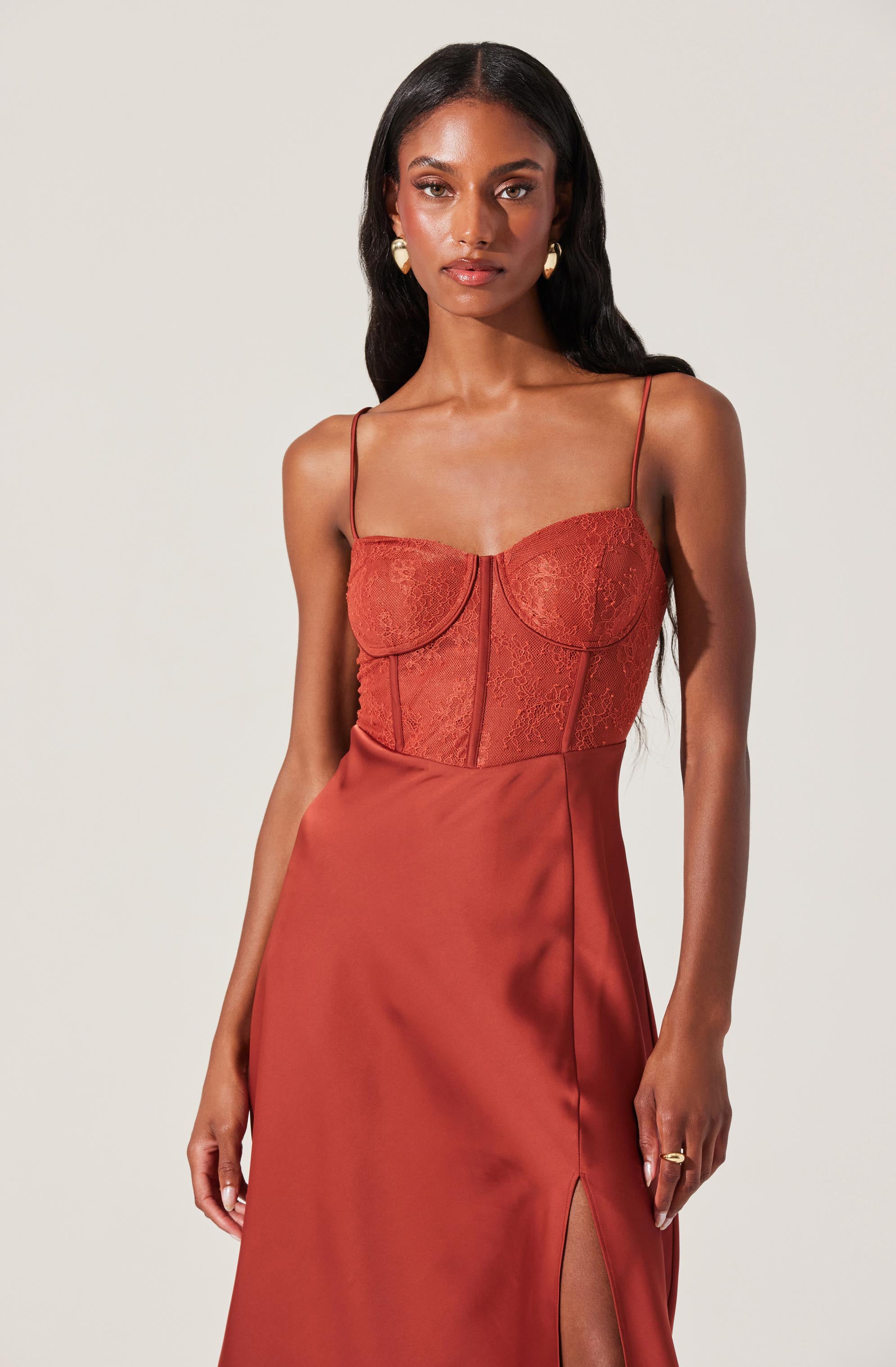 A woman is wearing a sleeveless, burnt orange dress with thin spaghetti straps. The bodice features delicate lace and structured seam details, while the skirt has a smooth satin finish with a slit on one side.