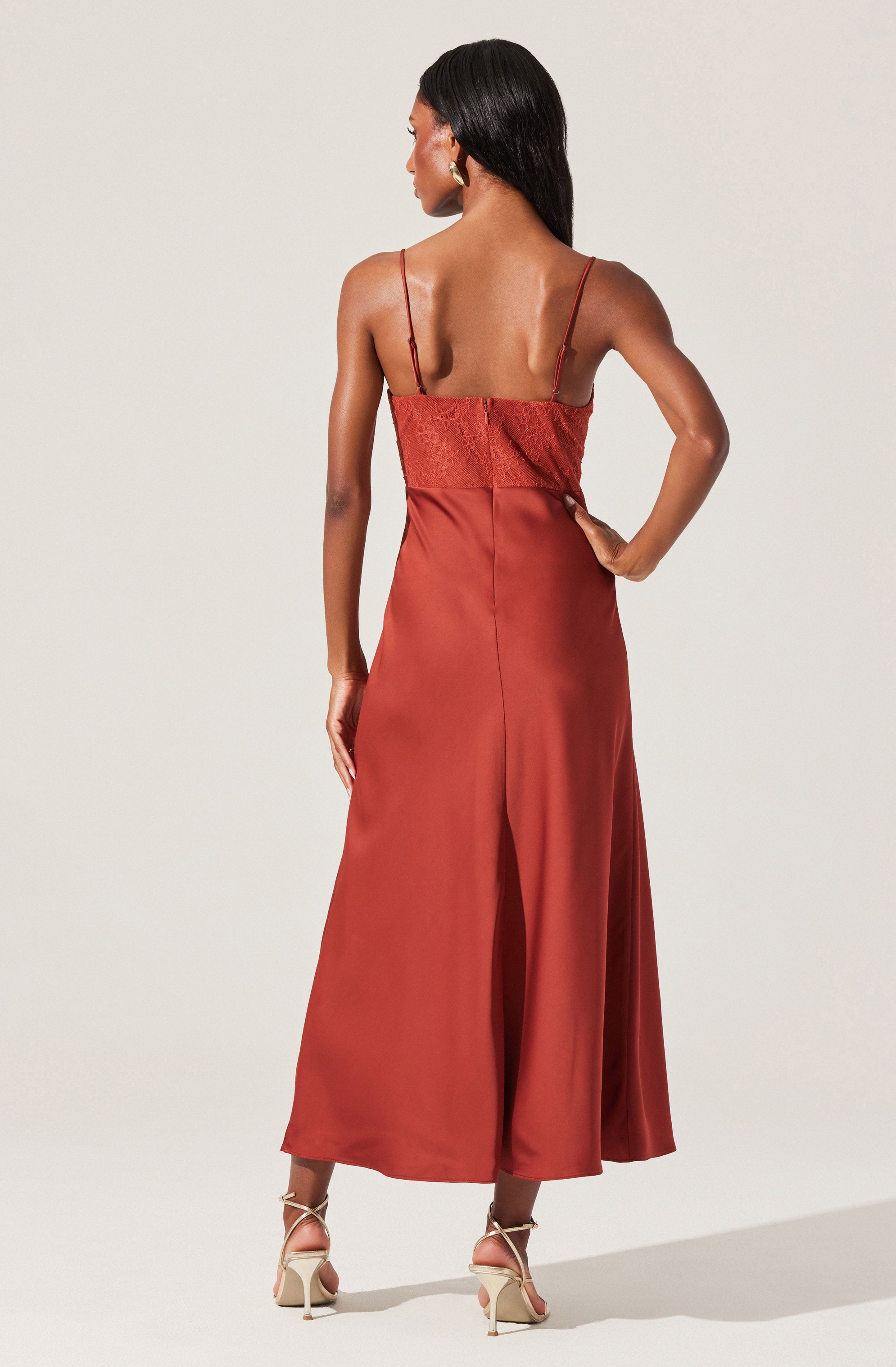 A woman is wearing a sleeveless, rust-colored, satin jumpsuit with thin spaghetti straps. The top features lace or embroidered detailing, and the bottom has wide-legged, cropped pants that reach just above the ankles. She is also wearing beige high-heeled sandals with ankle straps.
