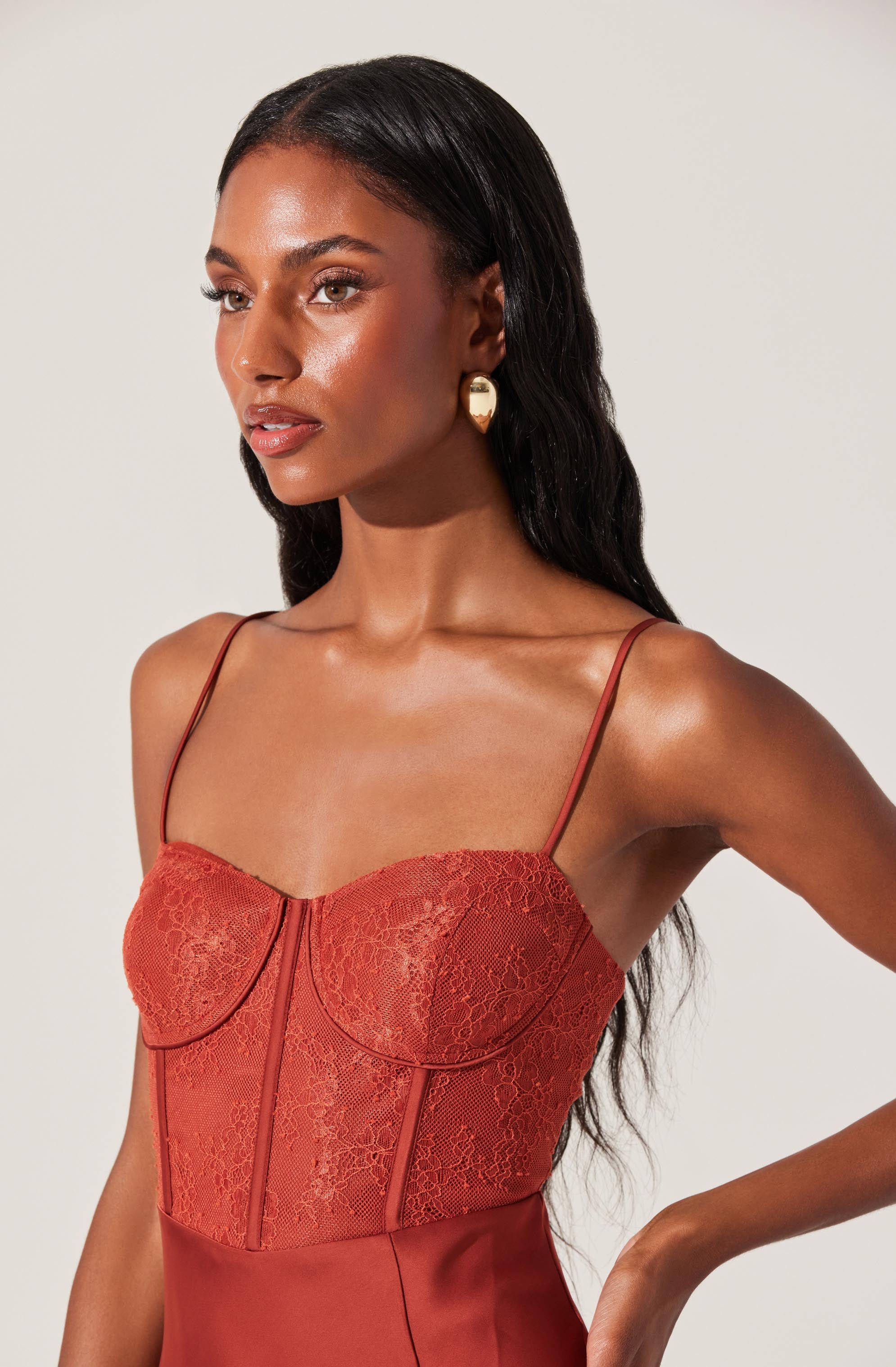 A woman is wearing a fitted dress with thin spaghetti straps. The bodice features a lace overlay with a floral pattern, and the lower part of the dress is made of smooth, satin-like fabric. The color of the dress is a warm rust or terracotta shade.
