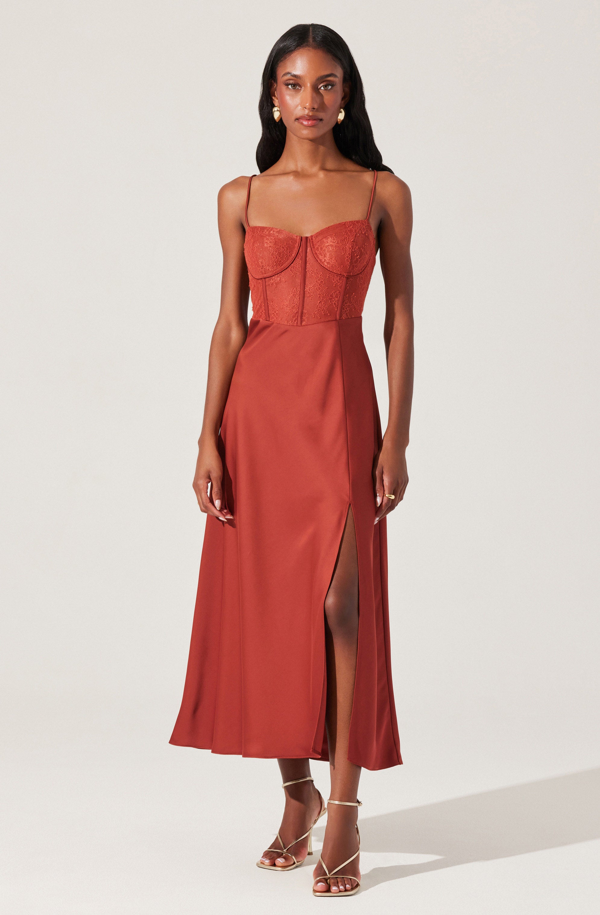 A woman is wearing a burnt orange, spaghetti strap maxi dress. The dress features a fitted bodice with a sweetheart neckline and lace detailing, and a high slit on one side revealing her leg. She is also wearing beige, strappy high-heeled sandals and gold statement earrings.