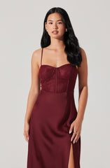 The woman is wearing a sleeveless, burgundy dress with thin spaghetti straps. The top part of the dress features a lace design with a sweetheart neckline and structured cups, while the lower part is smooth satin with a slit on one side.