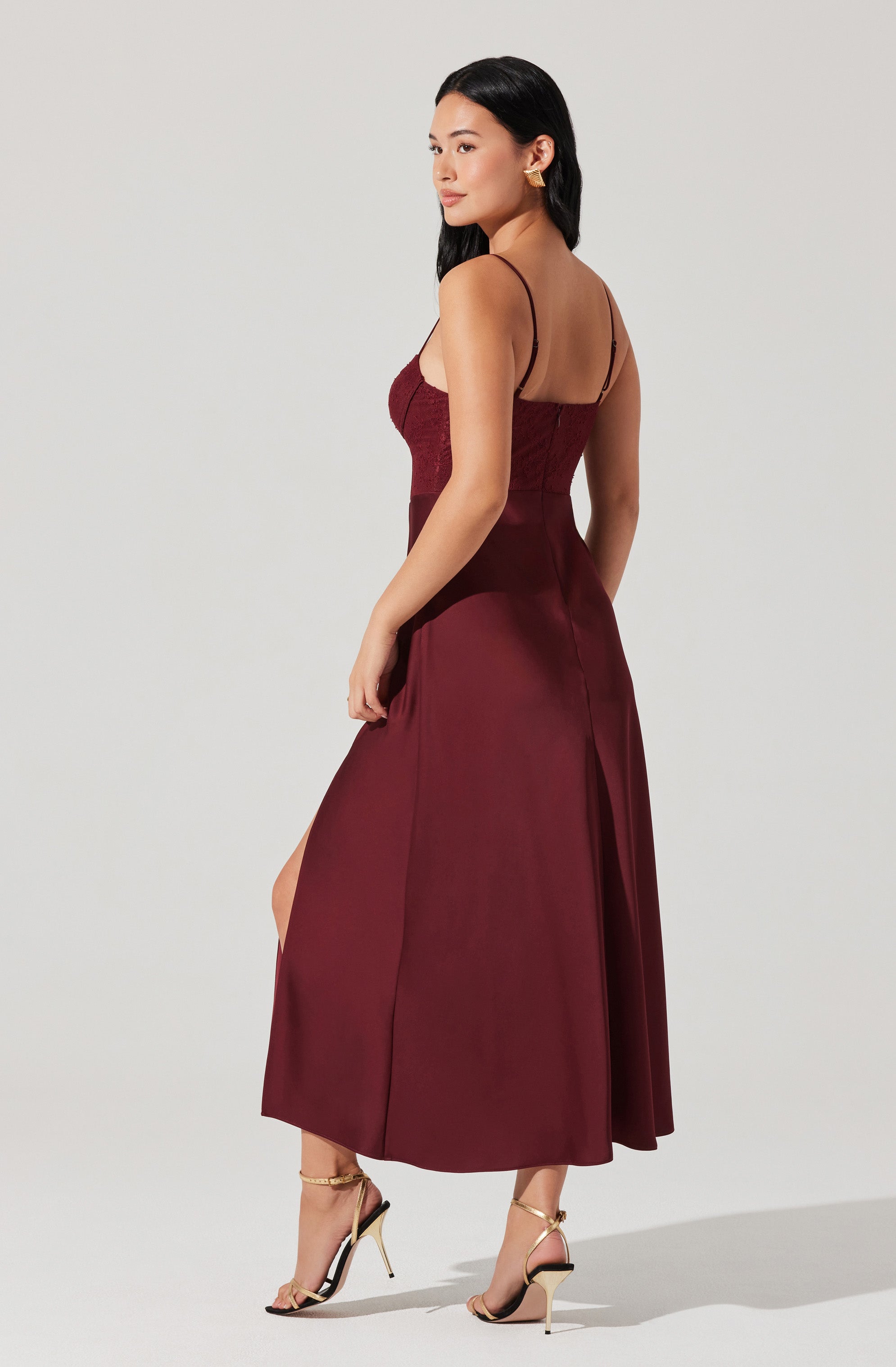 A woman is wearing a burgundy slip dress with thin spaghetti straps, fitted at the bodice with gathered fabric detailing, and a full-length, flowing skirt with a front slit. She is also wearing strappy high-heeled sandals with thin, gold ankle straps and stiletto heels.