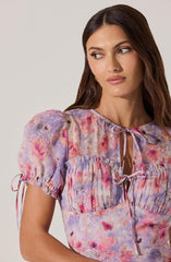 A woman is wearing a short-sleeved blouse with a watercolor floral pattern in shades of pink, purple, and peach. The blouse has gathered puff sleeves with ties at the cuffs, a ruffled yoke across the chest, and a front tie neckline.