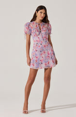 A woman is wearing a short, pastel-colored dress with a tie neckline and puffed sleeves. The dress features a watercolor-like pattern in shades of pink, purple, blue, and orange. She has on gold earrings and nude high-heeled sandals.