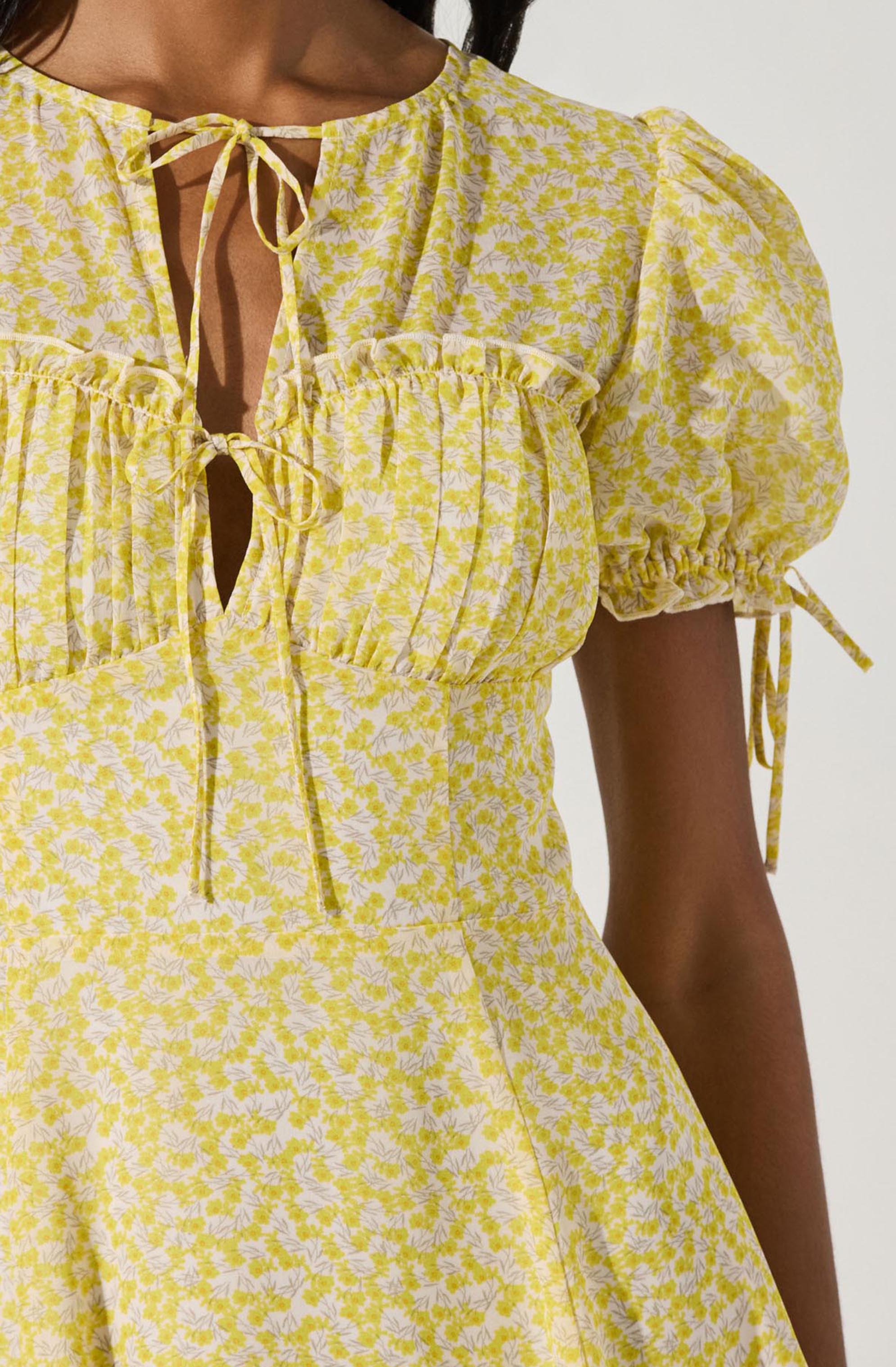 A close-up of a woman wearing a yellow dress with a white floral pattern. The dress features puffed short sleeves with ties at the ends, a gathered front with a small ruffle detail along the chest, and a keyhole neckline with adjustable ties. The fabric appears lightweight and textured.