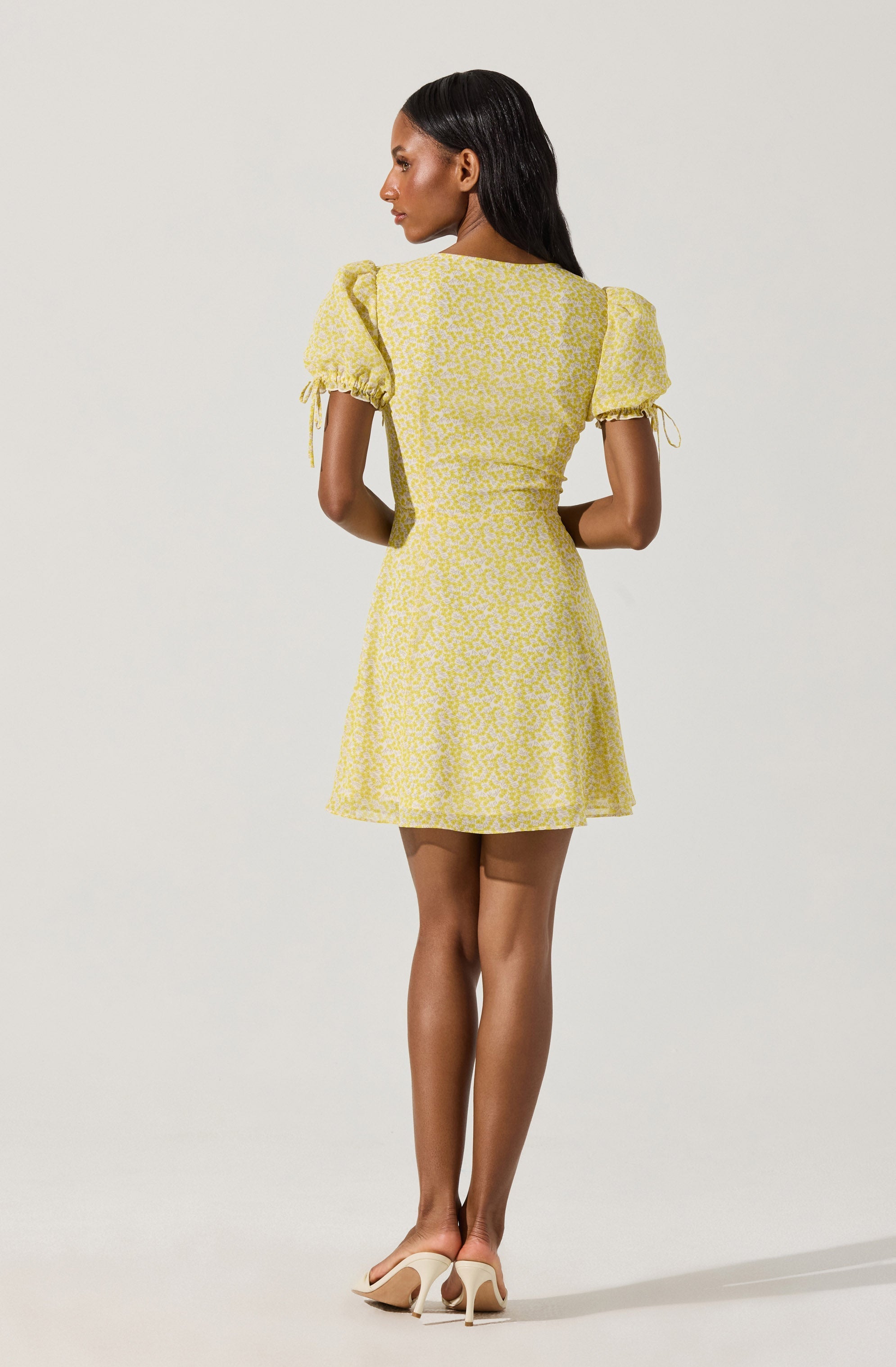 A woman is wearing a short, yellow dress with a white floral pattern. The dress has puffed short sleeves with ties at the ends, a fitted bodice, and a flared skirt. She is also wearing beige high-heeled shoes.