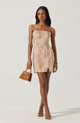 A woman is wearing a sleeveless, beige mini dress with a leopard print pattern and thin spaghetti straps. She accessorizes with a small tan handbag, gold jewelry including bracelets and rings, and nude high-heeled sandals.