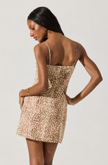 A woman is wearing a mini dress with a leopard print pattern. The dress has thin spaghetti straps and a fitted silhouette. She has dark, straight hair and is wearing a gold bracelet on her left wrist.