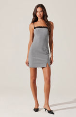 A woman is wearing a sleeveless, form-fitting gray dress with thin black spaghetti straps and a black lace trim along the top. The dress has a small slit at the front hem. She is also wearing black pointed-toe heels.