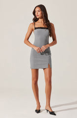 A woman is wearing a form-fitting, sleeveless mini dress that is predominantly gray with black lace trim at the neckline. The dress has a small slit on one side at the hem. She is also wearing black pointed-toe heels with a small heel and a bow detail on the front.