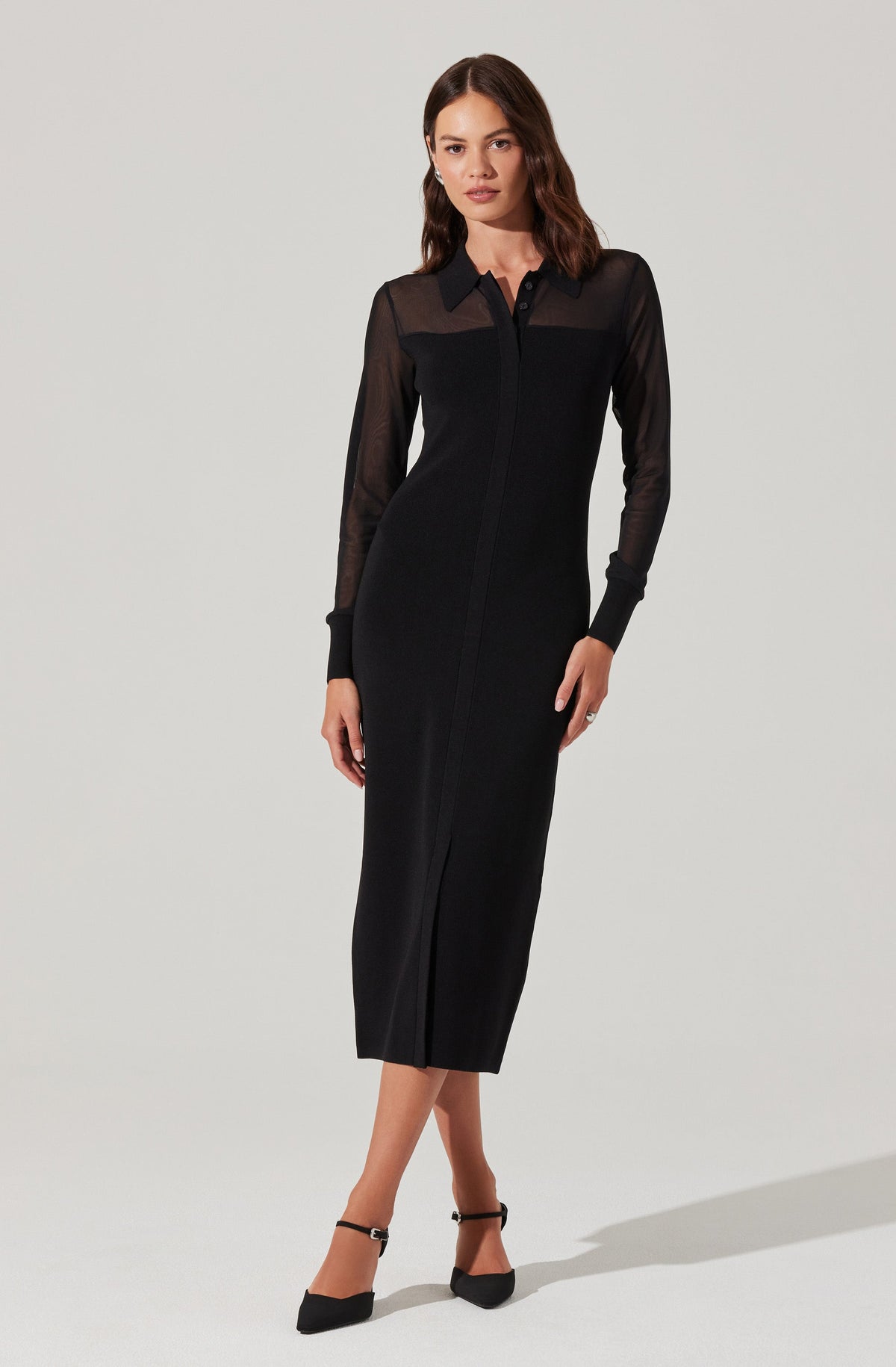 A woman is wearing a fitted, long black dress with a front zipper. The dress features sheer, long-sleeved panels on the upper chest, shoulders, and sleeves, contrasted by opaque fabric on the rest of the dress. She is also wearing black pointed-toe heels with ankle straps.