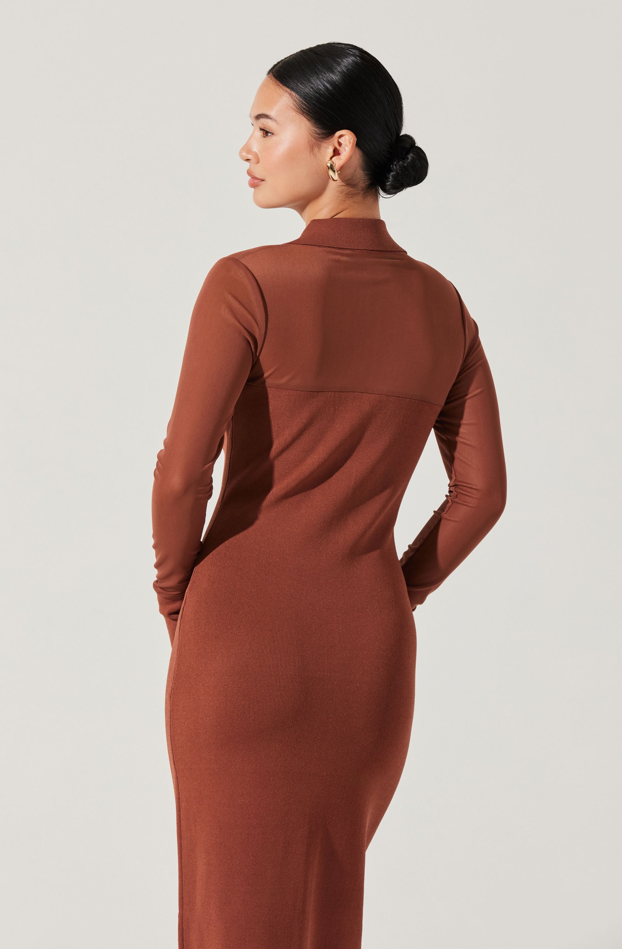 A woman is wearing a long, fitted dress in a warm brown color. The dress has a high neckline and long sleeves. The fabric appears smooth and slightly shiny, creating a sleek silhouette. The back of the dress does not have any visible embellishments or patterns. The woman has dark, straight hair styled in a low bun and is wearing gold hoop earrings.