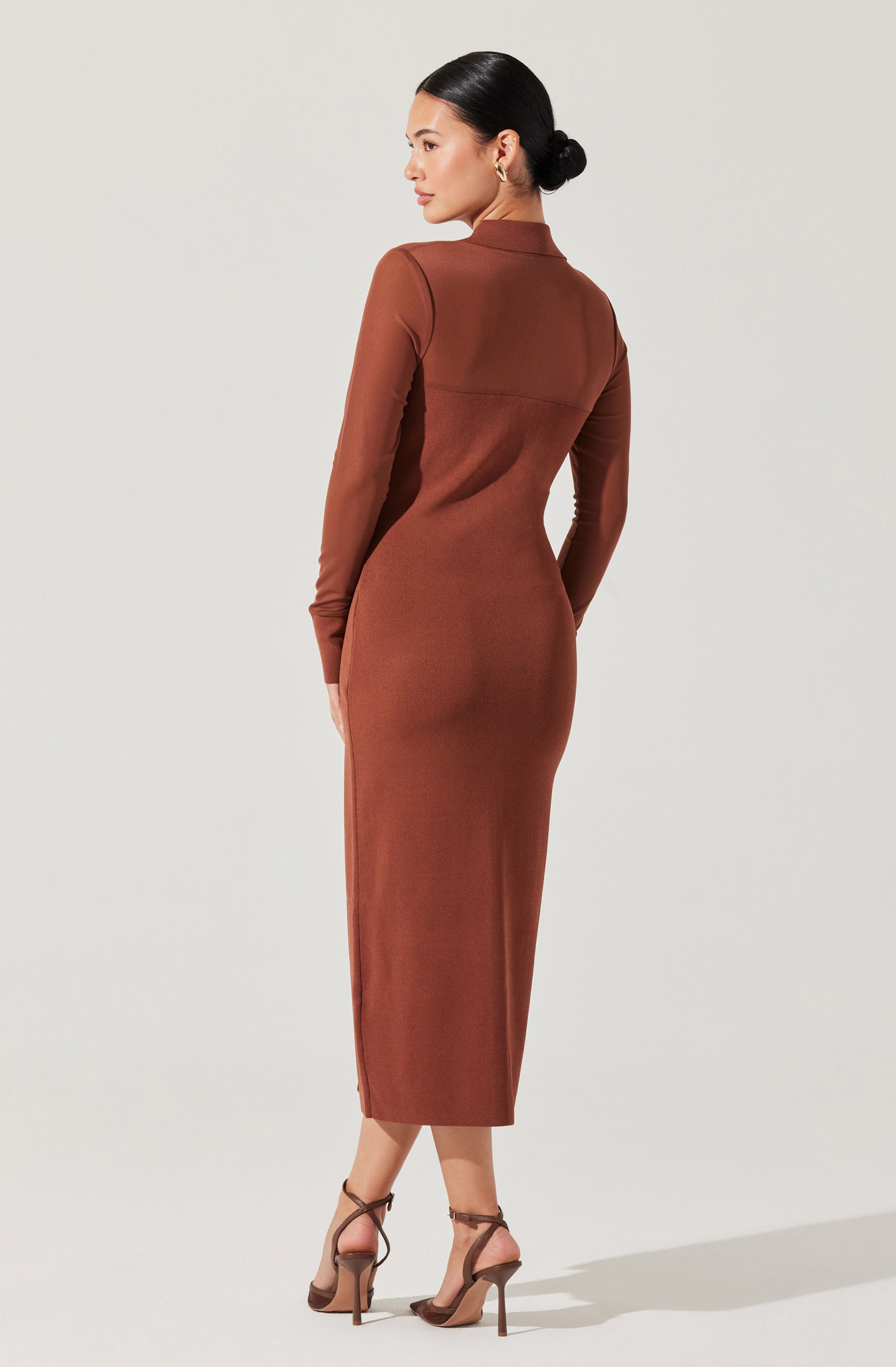 A woman is wearing a long, fitted, rust-colored dress with long sleeves and a high neckline. The dress has a seam detail across the upper back and falls to mid-calf length. She is pairing the dress with strappy high-heeled sandals. Her hair is styled in a sleek, low bun.