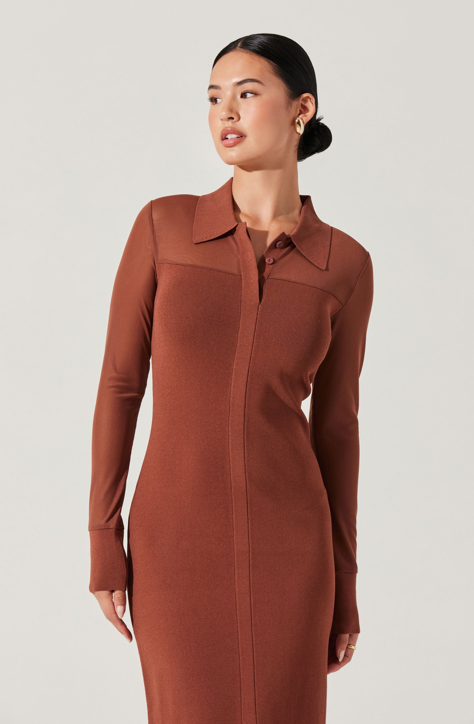 A woman is wearing a long-sleeved, fitted, rust-colored dress with a collared neckline and a front zipper. The dress is made of a smooth, structured fabric with a slightly textured panel across the chest and upper arms.