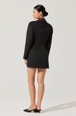 A woman is wearing a fitted black blazer-style dress with long sleeves and a collar. She has her hair styled in a low bun and is wearing earrings. The dress appears to be made of a structured fabric, creating a sleek and professional appearance. She is also wearing open-toe black heels.