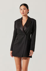 A woman is wearing a tailored black blazer dress with a satin lapel collar and long sleeves. The dress has a single button closure at the waist and features two flap pockets on the front.
