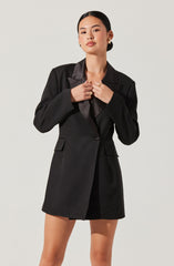 The woman is wearing a tailored black blazer with a shawl collar made of shiny satin fabric, paired with black shorts. She has gold hoop earrings and a gold ring, and she is posing with her hands near her neck.