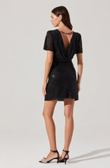 A woman is wearing a black, short-sleeved dress that is covered in shiny sequins. The dress features a cowl neckline at the back with a thin strap across the shoulders. She is also wearing black high-heeled sandals with ankle straps and decorative bows on the toes.
