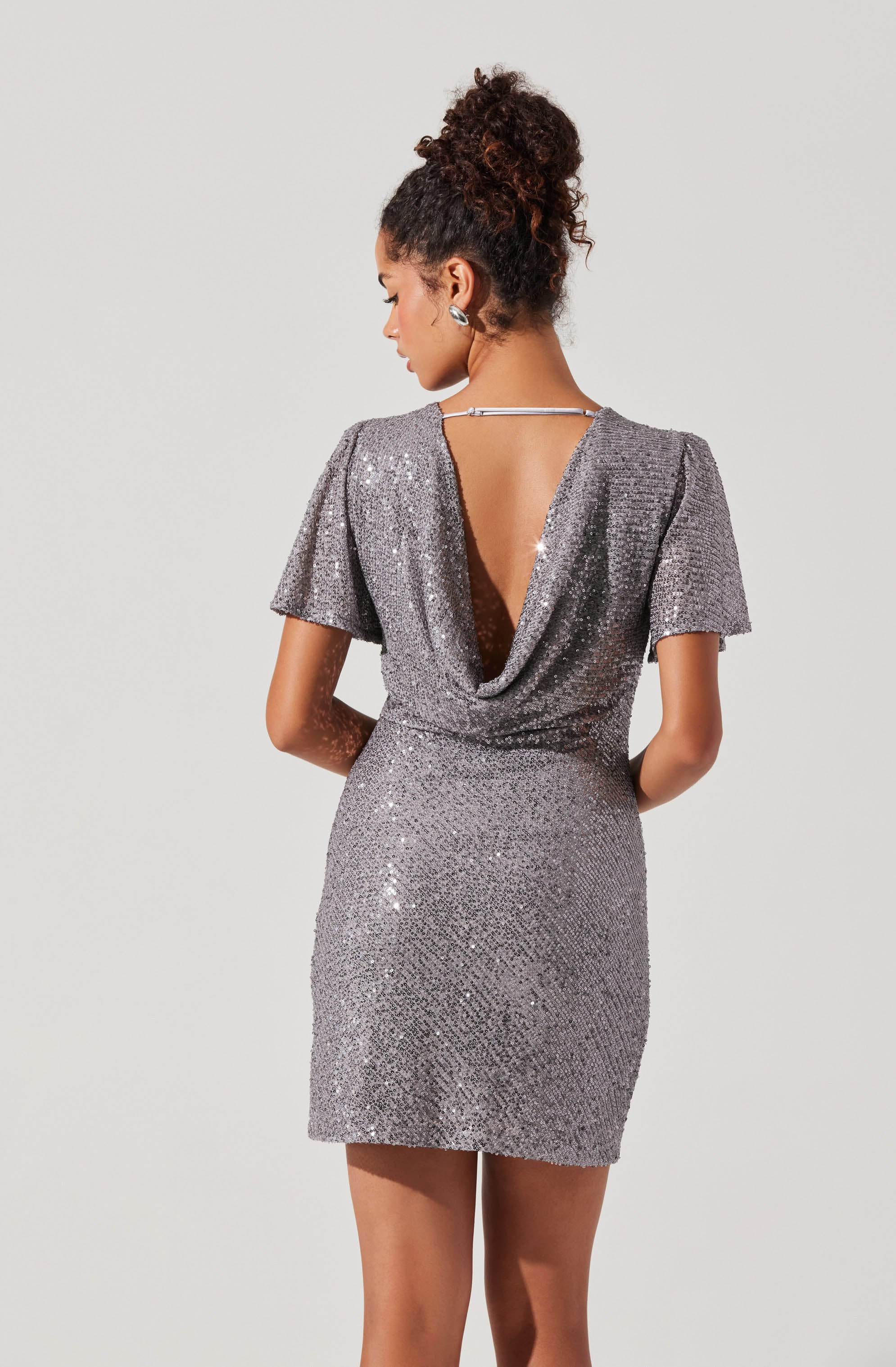 A woman is wearing a short, silver, sequined dress with a deep back neckline that features a draped cowl design. The dress has short, slightly puffed sleeves and is fitted at the waist, ending above the knees. She has her hair styled in a curly updo.