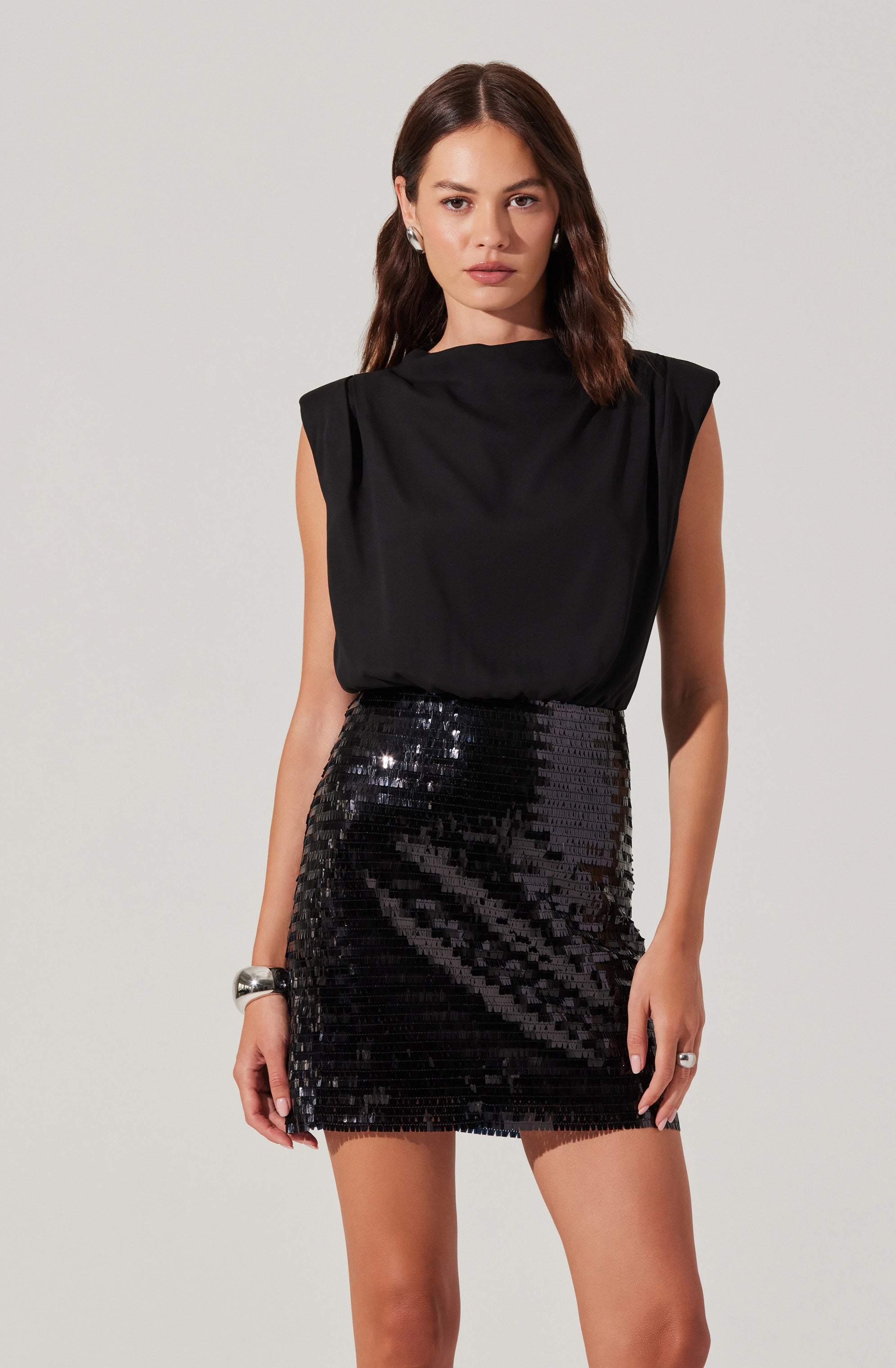 A woman is wearing a sleeveless black top with padded shoulders and a high neckline. She pairs it with a black sequined mini skirt. She has her hair styled in loose waves and is accessorizing with earrings, a bracelet, and a ring.