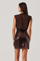 A woman is wearing a sleeveless, dark brown top with wide shoulder pads and a keyhole opening at the back. She is also wearing a high-waisted, short, fitted skirt covered in dark, shiny sequins that reflect light.