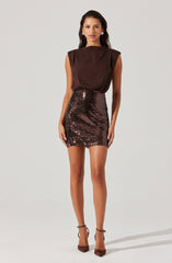 The woman is wearing a sleeveless, dark brown satin top with padded shoulders and a high neckline. She pairs it with a high-waisted, fitted mini skirt covered in shiny, dark sequins. She completes her outfit with dark brown high-heeled shoes that have ankle straps.