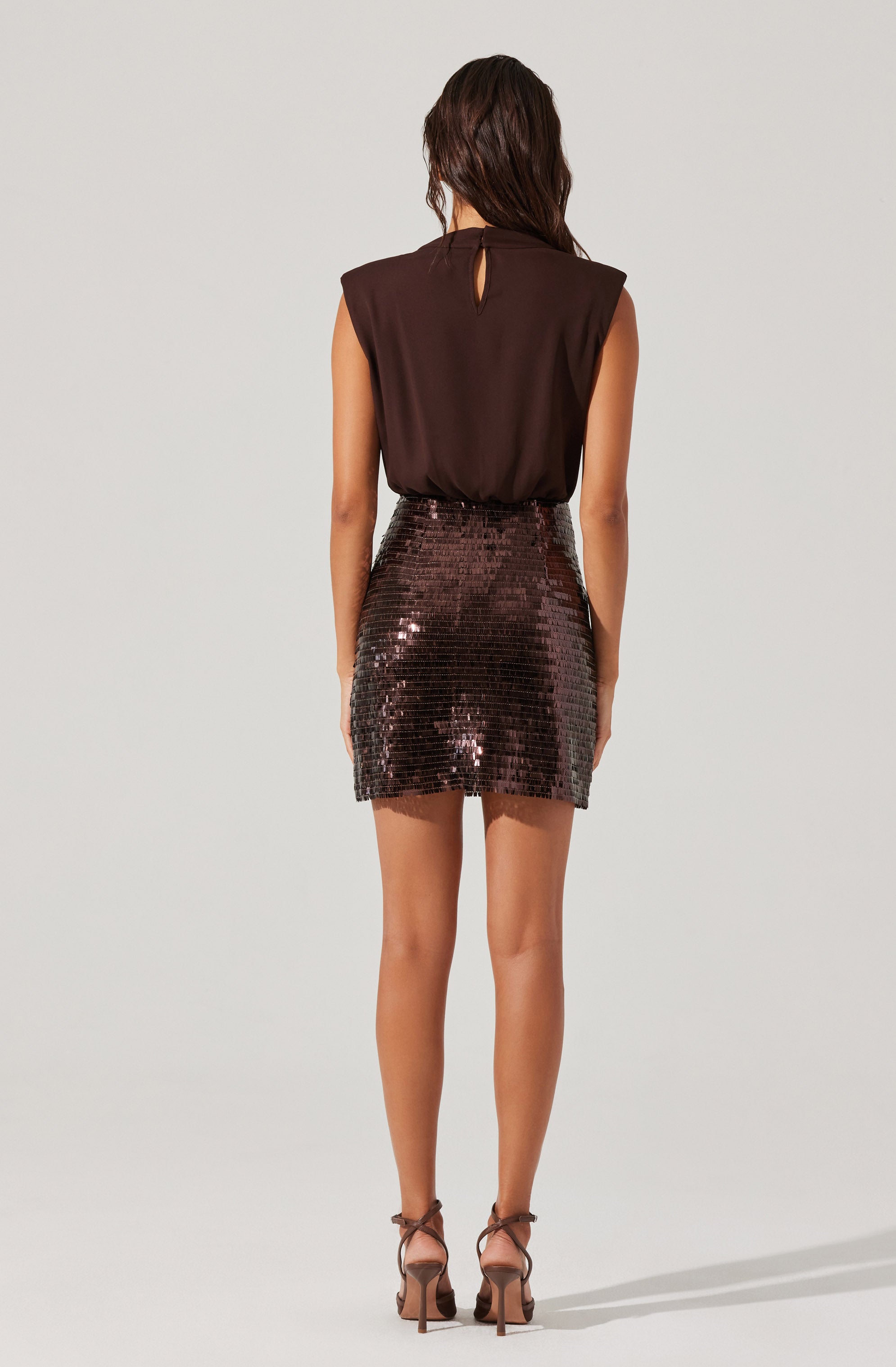 A woman is wearing a sleeveless dark brown top with a small keyhole opening at the back neckline, paired with a high-waisted, short skirt covered in shiny, dark sequins. She is also wearing strappy high-heeled sandals.