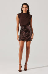 A woman is wearing a sleeveless, dark brown satin top with padded shoulders and a high neckline, paired with a high-waisted, fitted mini skirt covered in shiny, sequined fabric with a dark brown hue. She is also wearing dark brown ankle-strap high heels.