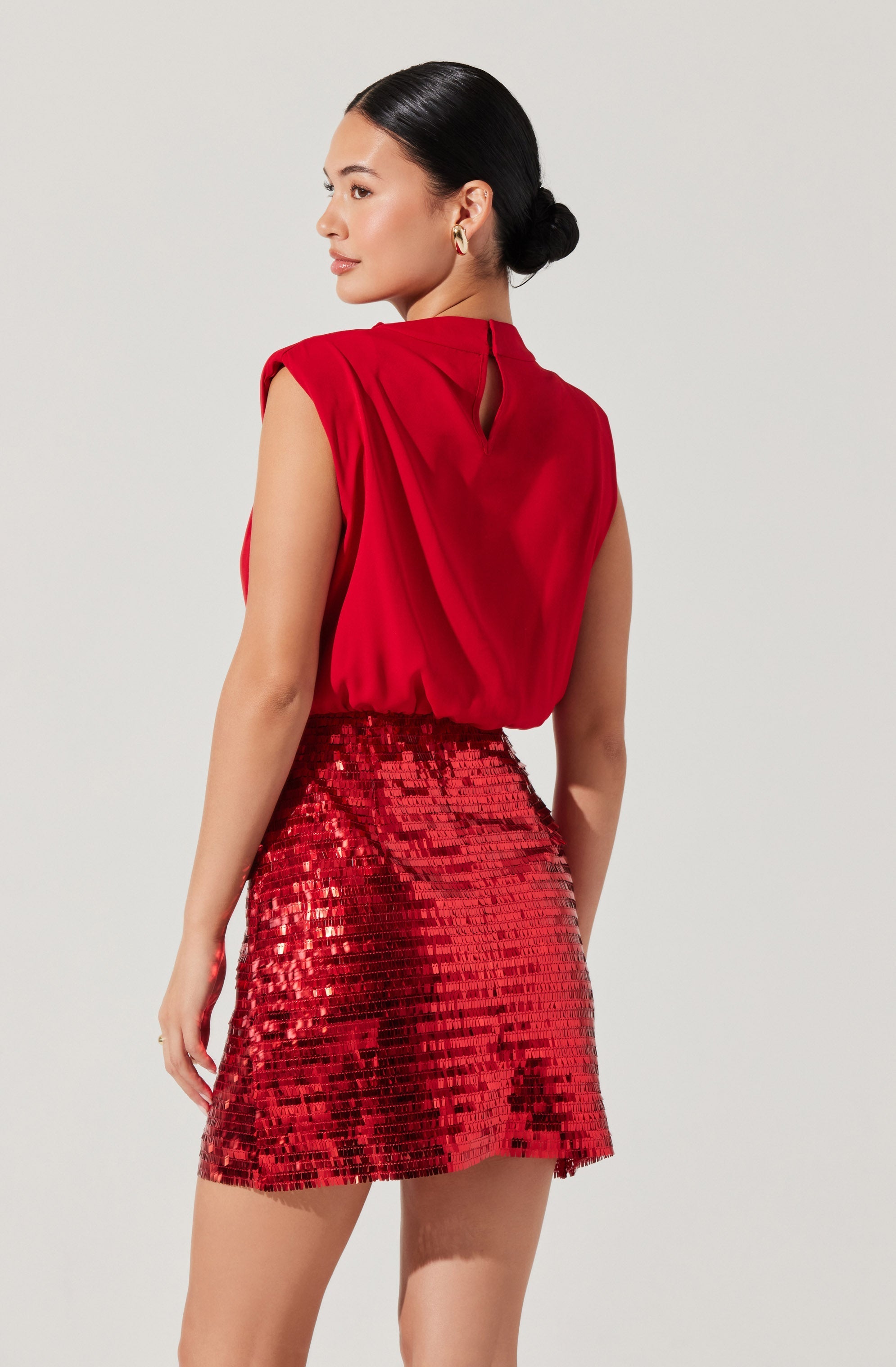 A woman is wearing a sleeveless, red blouse with a small keyhole opening at the back, paired with a high-waisted, red sequined mini skirt.