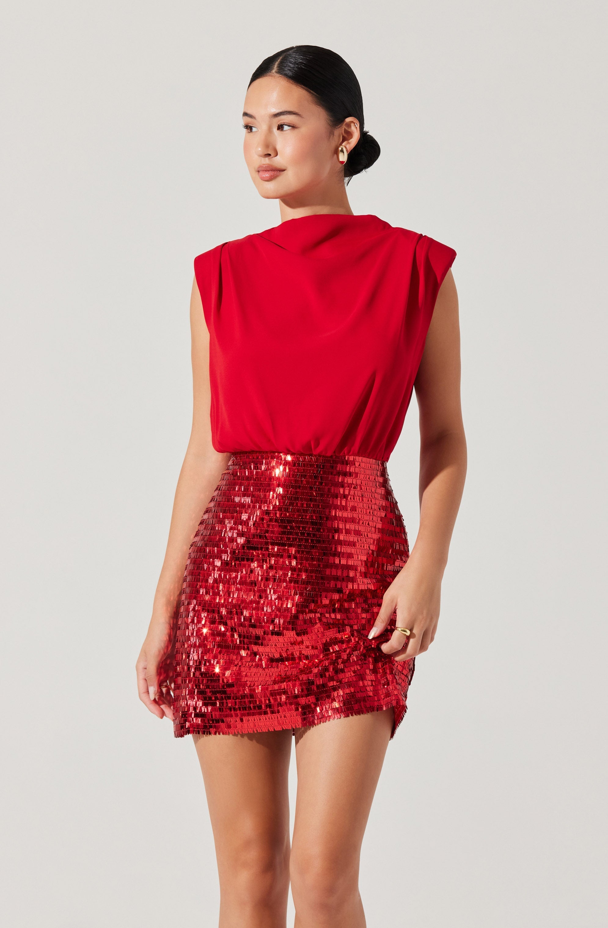 A woman is wearing a sleeveless red top with a draped neckline and padded shoulders, paired with a high-waisted, fitted mini skirt covered in shiny red sequins.