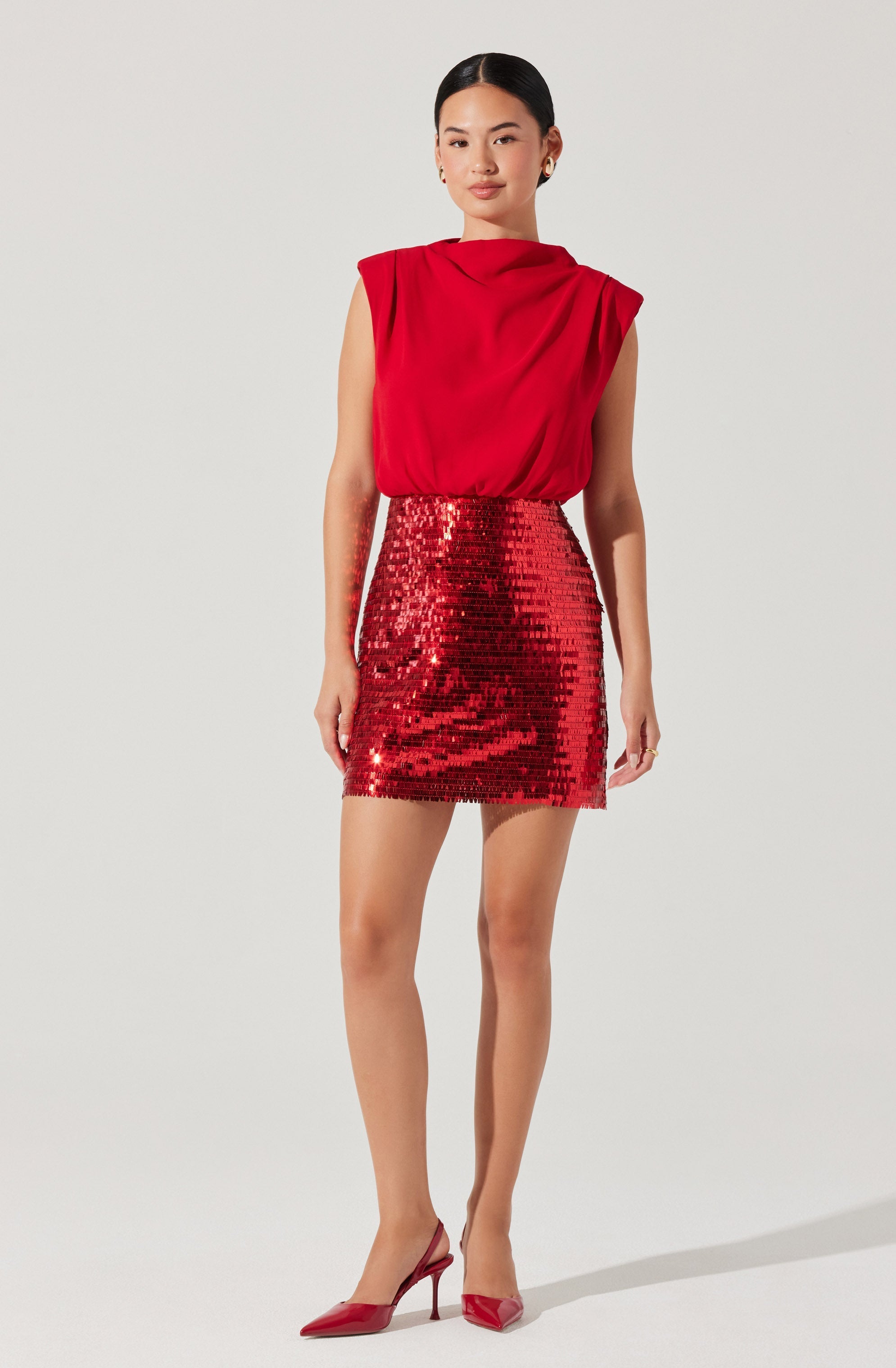 A woman is wearing a sleeveless red top with a draped neckline and padded shoulders. She pairs it with a high-waisted, fitted mini skirt covered in shiny red sequins. She completes the outfit with red pointed-toe high-heeled shoes.