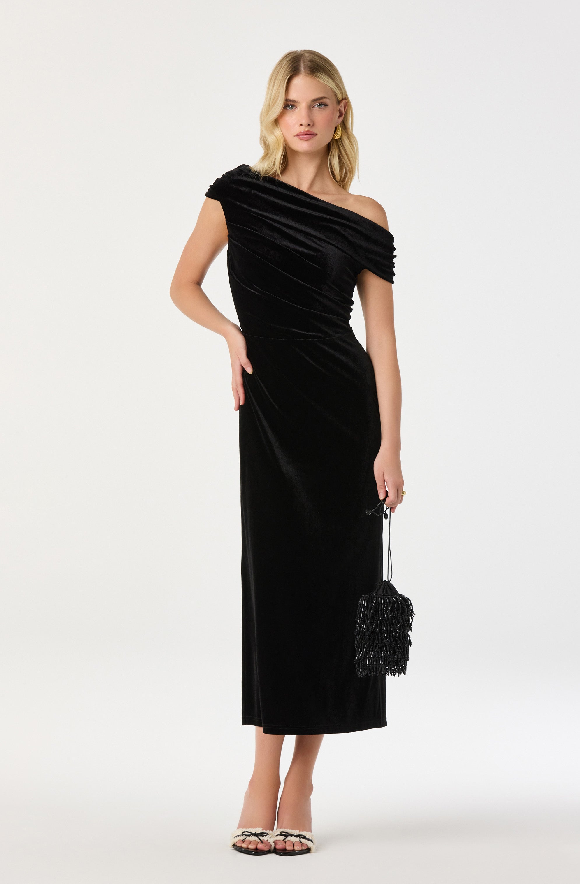 A woman is wearing a black, one-shoulder, floor-length velvet evening gown with a ruched bodice. She is holding a small black fringe handbag in her right hand and has minimal accessories, including earrings. She is also wearing open-toe heels with a black and white pattern.