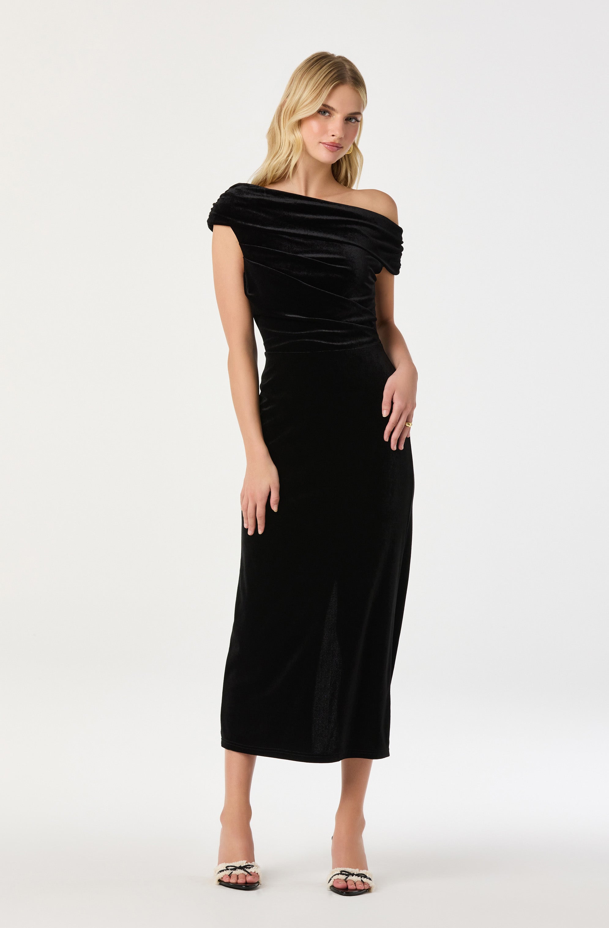 A woman is wearing an elegant black off-the-shoulder evening gown with a fitted waist and a long, flowing skirt. The top part of the dress features gathered fabric that creates a textured, draped effect around the neckline and shoulders. She is also wearing open-toed shoes with a black and white pattern.