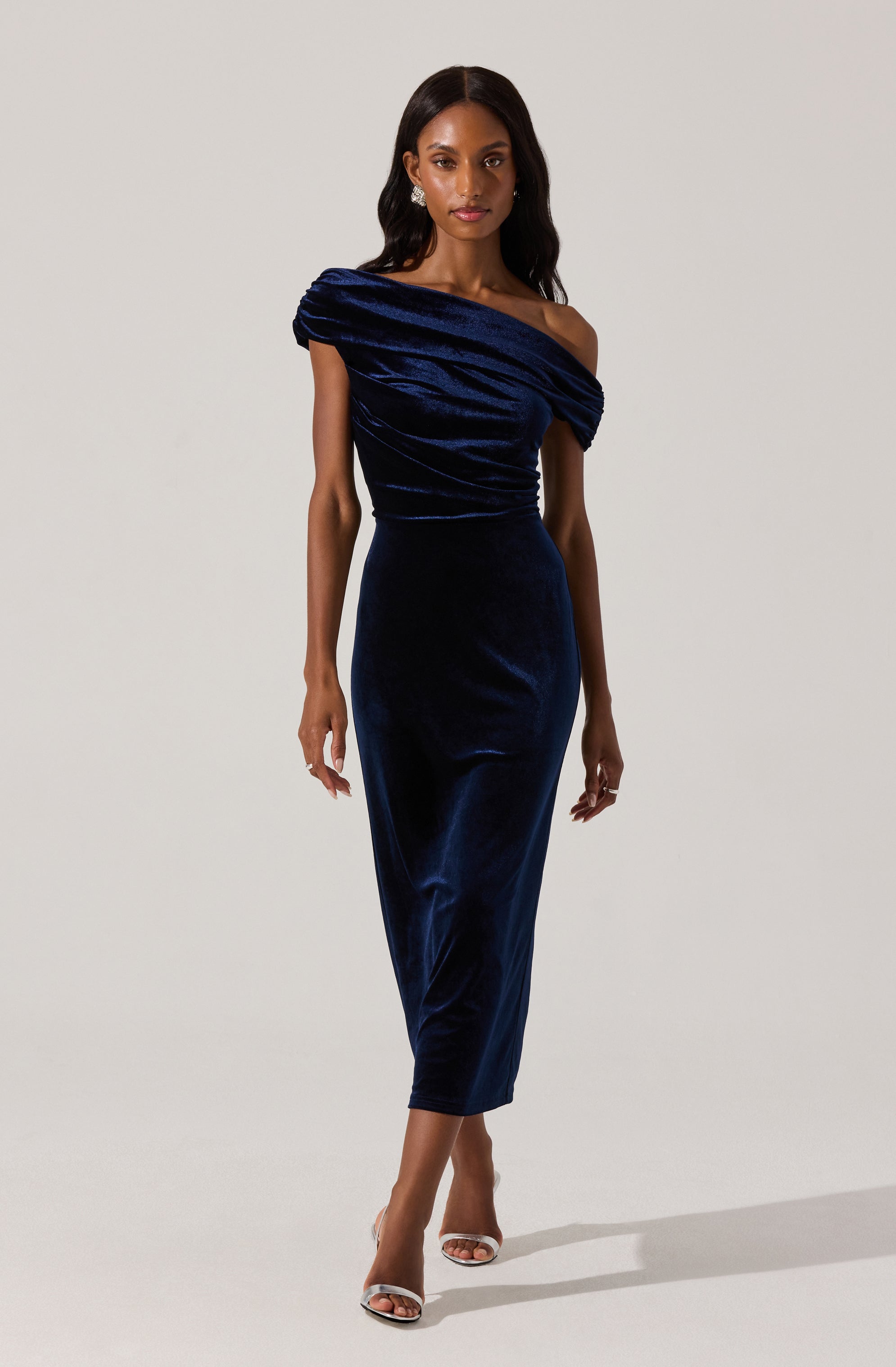 A woman is wearing a dark blue velvet dress with an off-the-shoulder, draped neckline on one side. The dress is fitted and long, reaching down to her ankles. She is also wearing open-toed high heels with a light-colored strap.