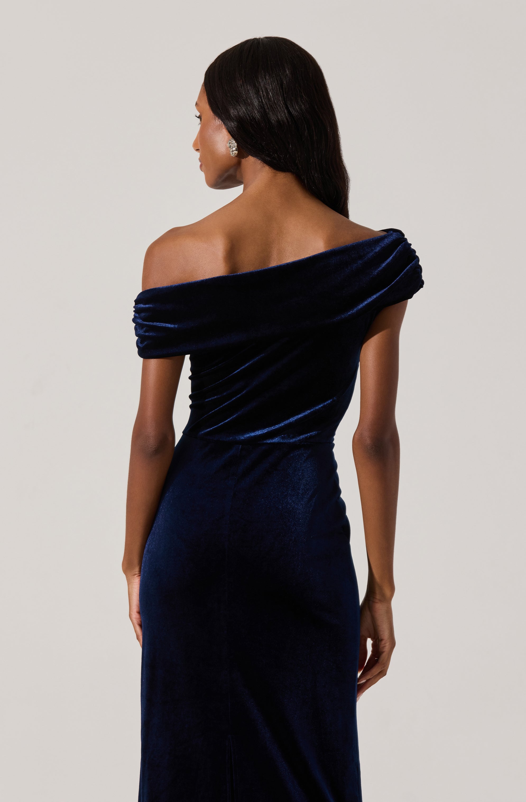 A woman is wearing an elegant off-the-shoulder evening gown made of deep navy blue velvet fabric. The dress features a fitted silhouette that accentuates her figure, with gathered detailing around the neckline and shoulders, creating a soft draped effect. The fabric has a rich, plush texture and a subtle sheen.
