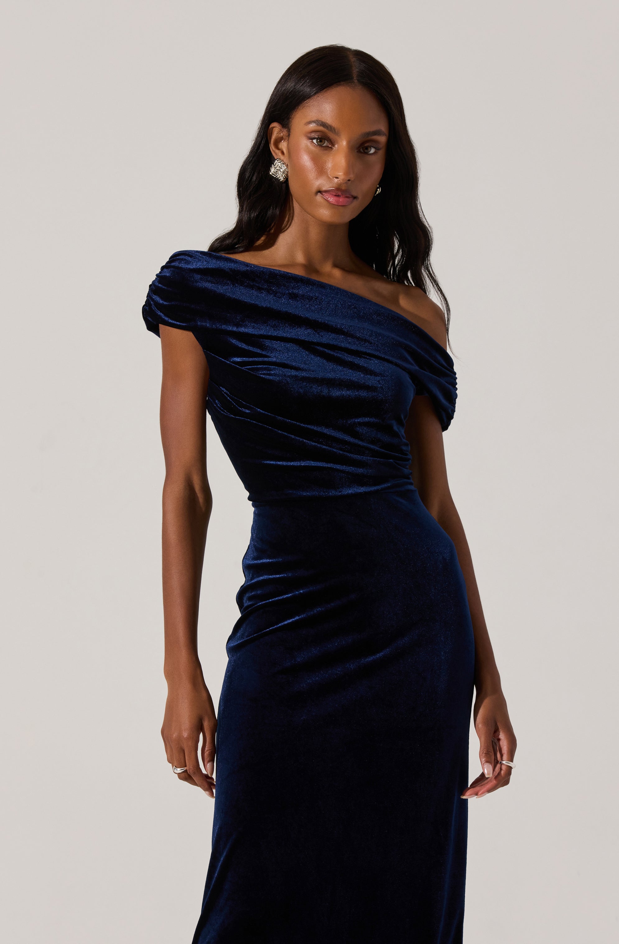 A woman is wearing a dark blue, velvet, off-the-shoulder gown with gathered fabric at the bodice and sleeves, creating a ruched appearance.
