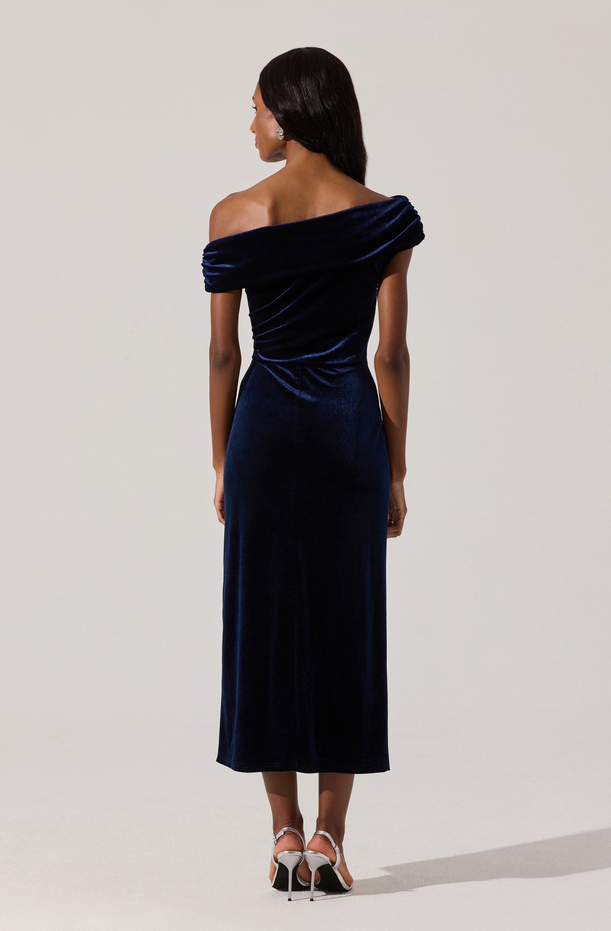 A woman is wearing a dark blue velvet off-the-shoulder gown with a draped neckline and long length. She is also wearing white high-heeled sandals.