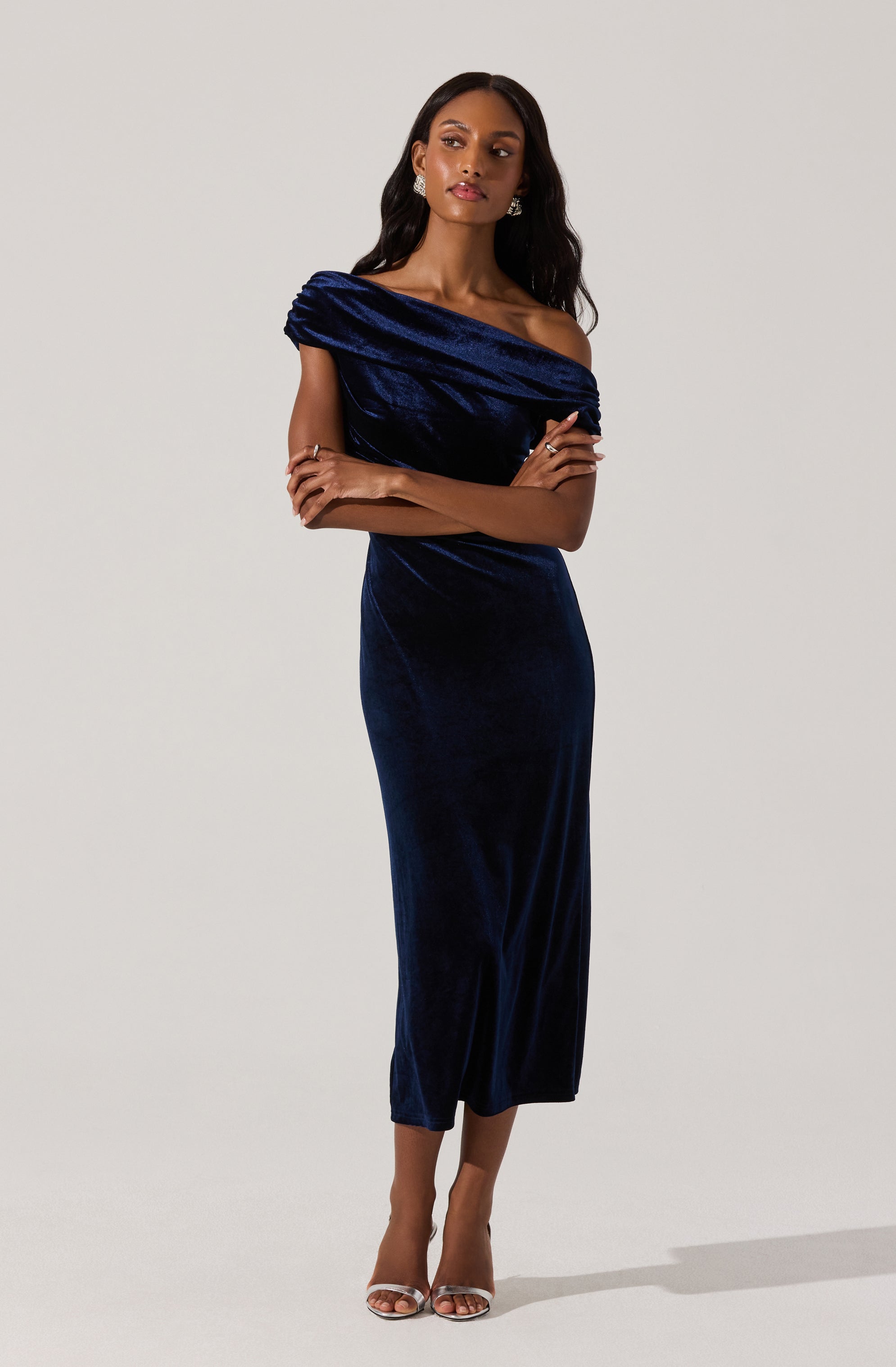 A woman is wearing a dark blue velvet dress that features an off-shoulder design with a draped, gathered bodice style. The dress has a fitted silhouette that extends to a midi length, ending mid-calf. She is also wearing silver open-toe heels and sparkling earrings.