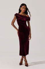 A woman is wearing a deep burgundy velvet gown with an off-the-shoulder neckline and draped, gathered fabric at the top. The dress is fitted at the waist and extends to a midi length, reaching just above her ankles. She is paired with matching burgundy heels with ankle straps.