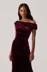 A woman is wearing an elegant, off-the-shoulder evening gown made of deep burgundy velvet fabric. The dress features draped, ruched detailing across the bust and fitted silhouette that emphasizes her figure. The fabric has a soft sheen, adding a luxurious touch to the overall look.