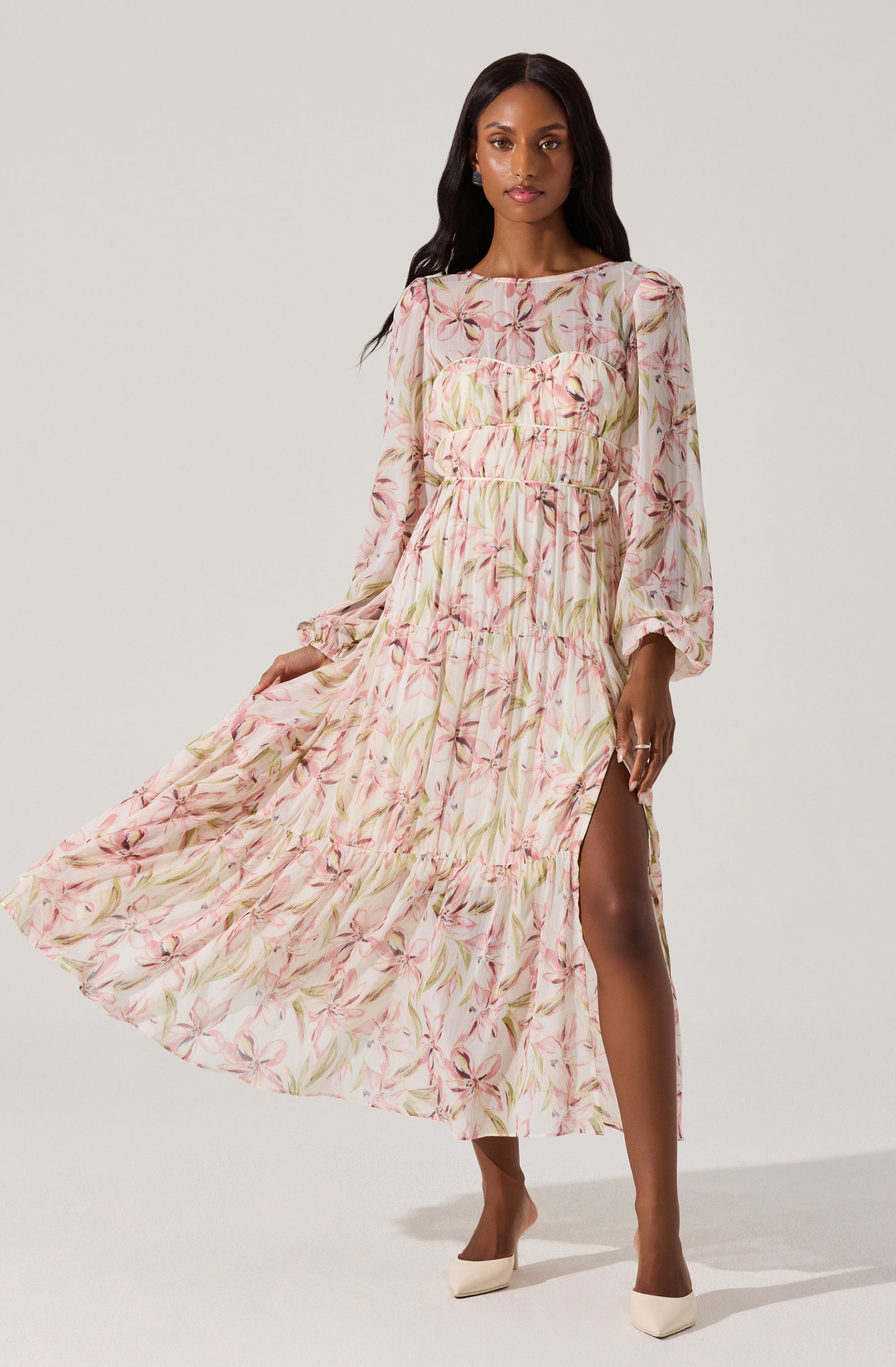 A woman is wearing a long, flowing dress with a light, pastel-colored floral print. The dress features long sleeves with elastic cuffs, a round neckline, and a tiered, ruffled skirt with a high slit on one side. The fabric appears lightweight and semi-sheer, with a soft pink and beige color palette accented by green leaves and pinkish flowers. She pairs the dress with beige high heels.