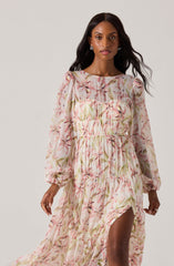 A woman is wearing a long, flowing dress with a floral pattern that features pink, green, and beige colors. The dress has long puffed sleeves with elastic cuffs and a ruffled slit that reveals her leg. The fabric appears lightweight and semi-sheer, with a gathered and tiered design creating a textured look.