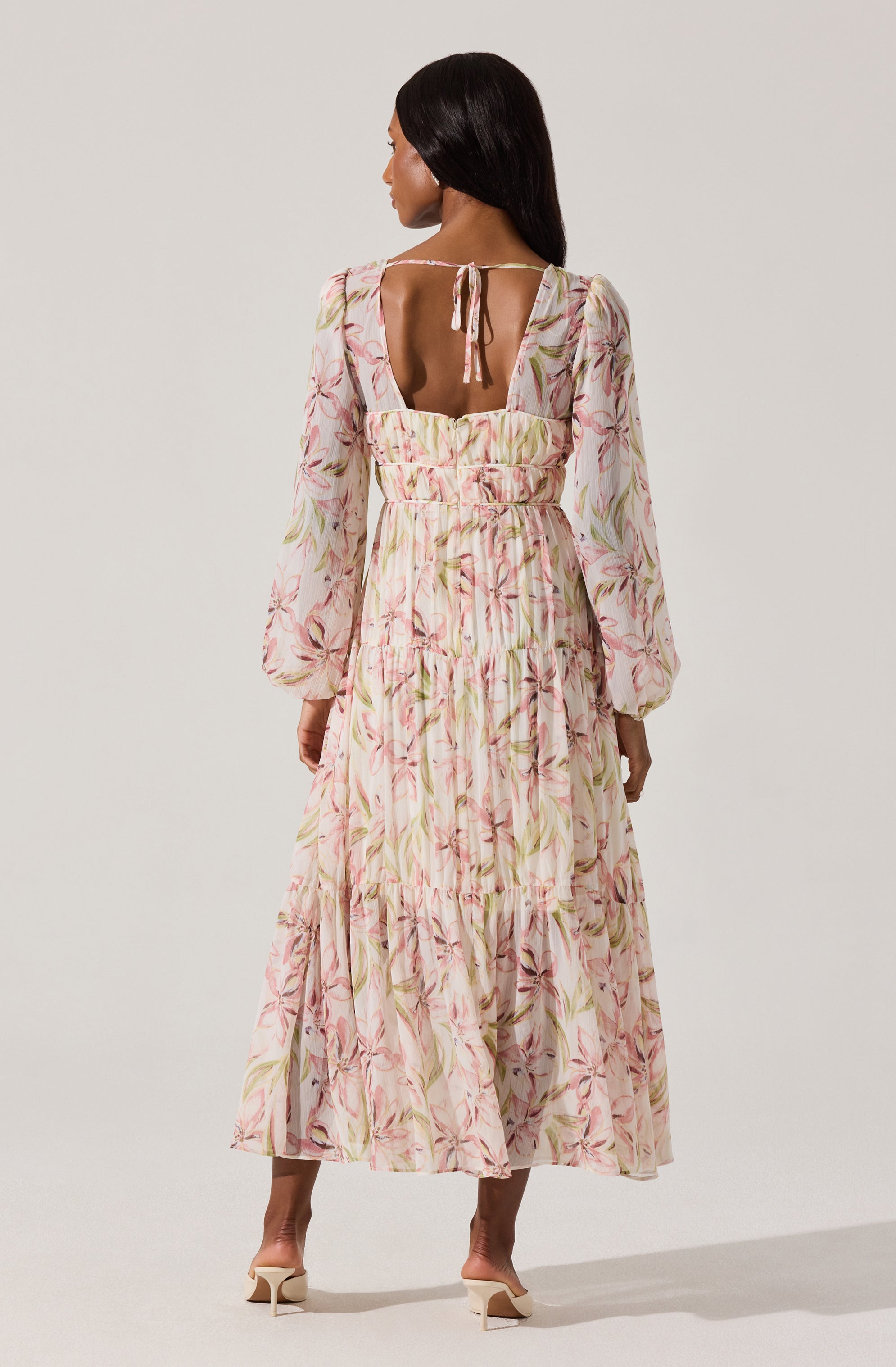 A woman is wearing a long, flowy dress with a light background and a soft pink, green, and beige floral pattern. The dress features puffy long sleeves, a fitted waist, and a tiered skirt. The back of the dress has a square cutout with a small bow tied at the top of the opening.