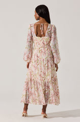 A woman is wearing a long, flowy dress with a light background and a soft pink, green, and beige floral pattern. The dress features puffy long sleeves, a fitted waist, and a tiered skirt. The back of the dress has a square cutout with a small bow tied at the top of the opening.