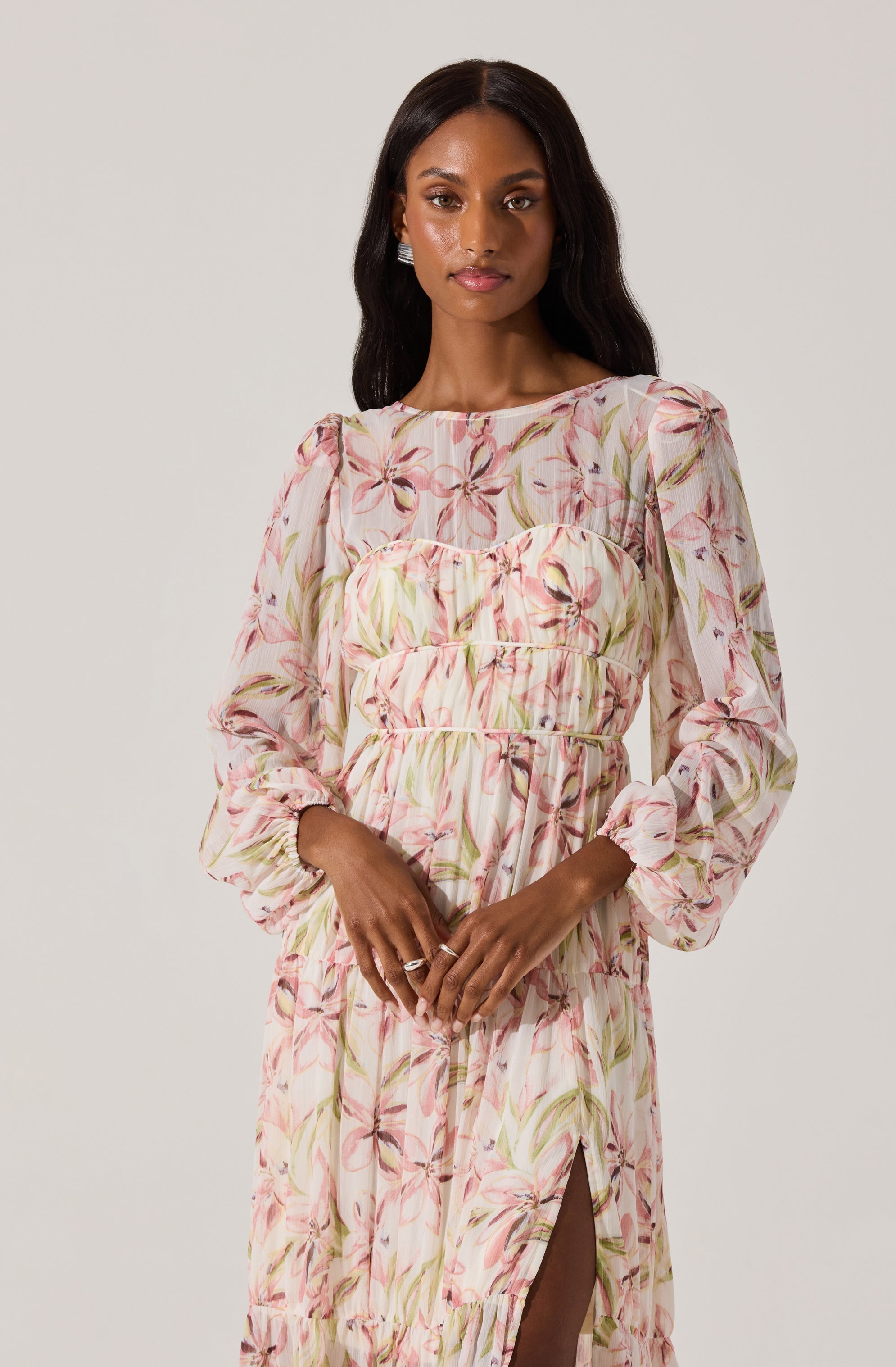 A woman is wearing a long, flowing dress with a pastel floral pattern. The dress has long, puffed sleeves that are gathered at the cuffs, and a high slit on one side revealing her leg. The fabric features soft pink and green flowers with light-colored leaves.